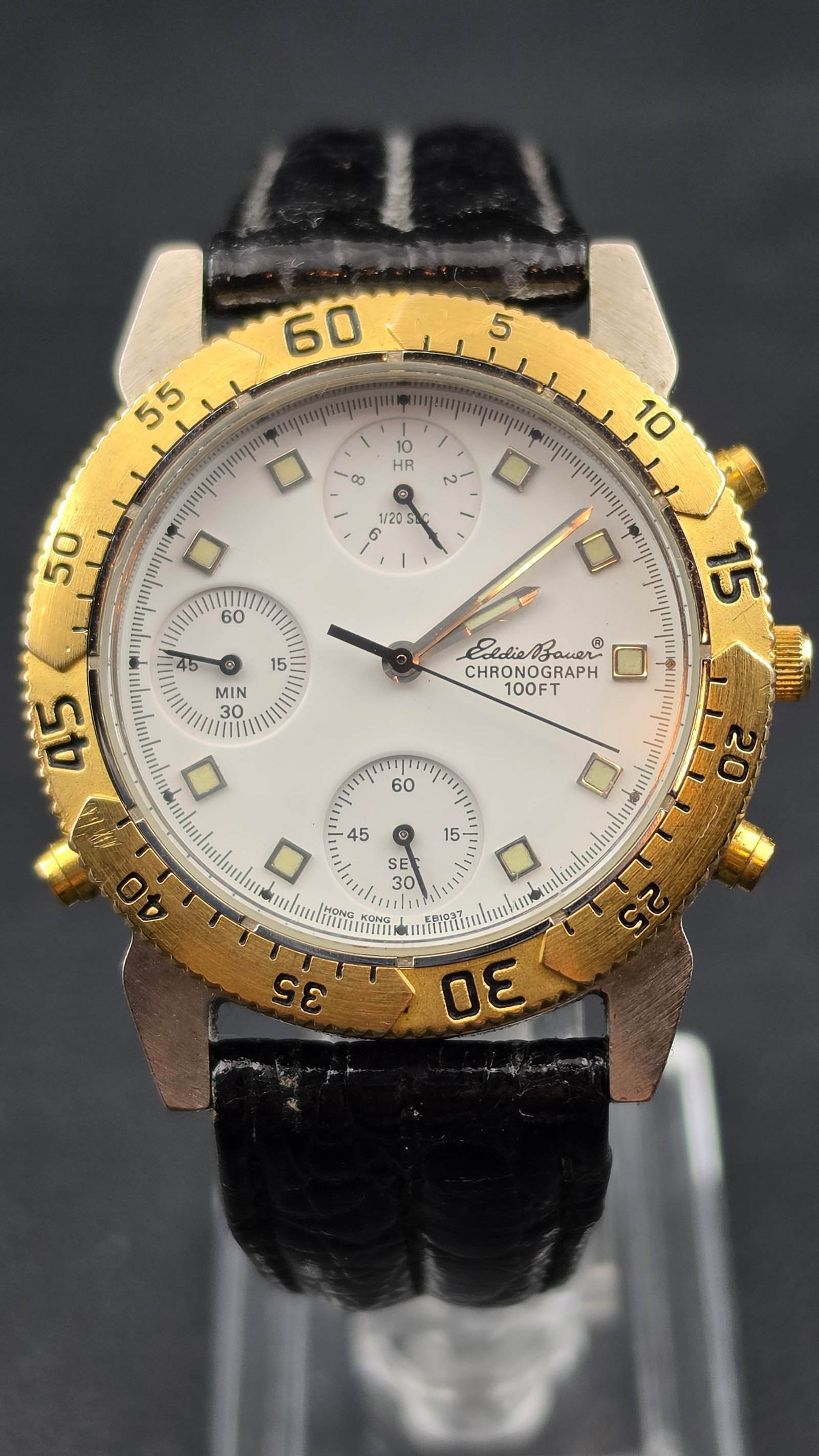 Eddie Bauer Chronograph 100FT Quartz Watch 38.17mm Gold Tone Bezel EB1037 219-0333: White dial with Eddie Bauer script text, Chronograph and 100FT text, three subdials including a 10-hour and 1/20 sec counter at 12, a minutes counter at 9 showing 60, 45, 15, and 30, and a seconds cou