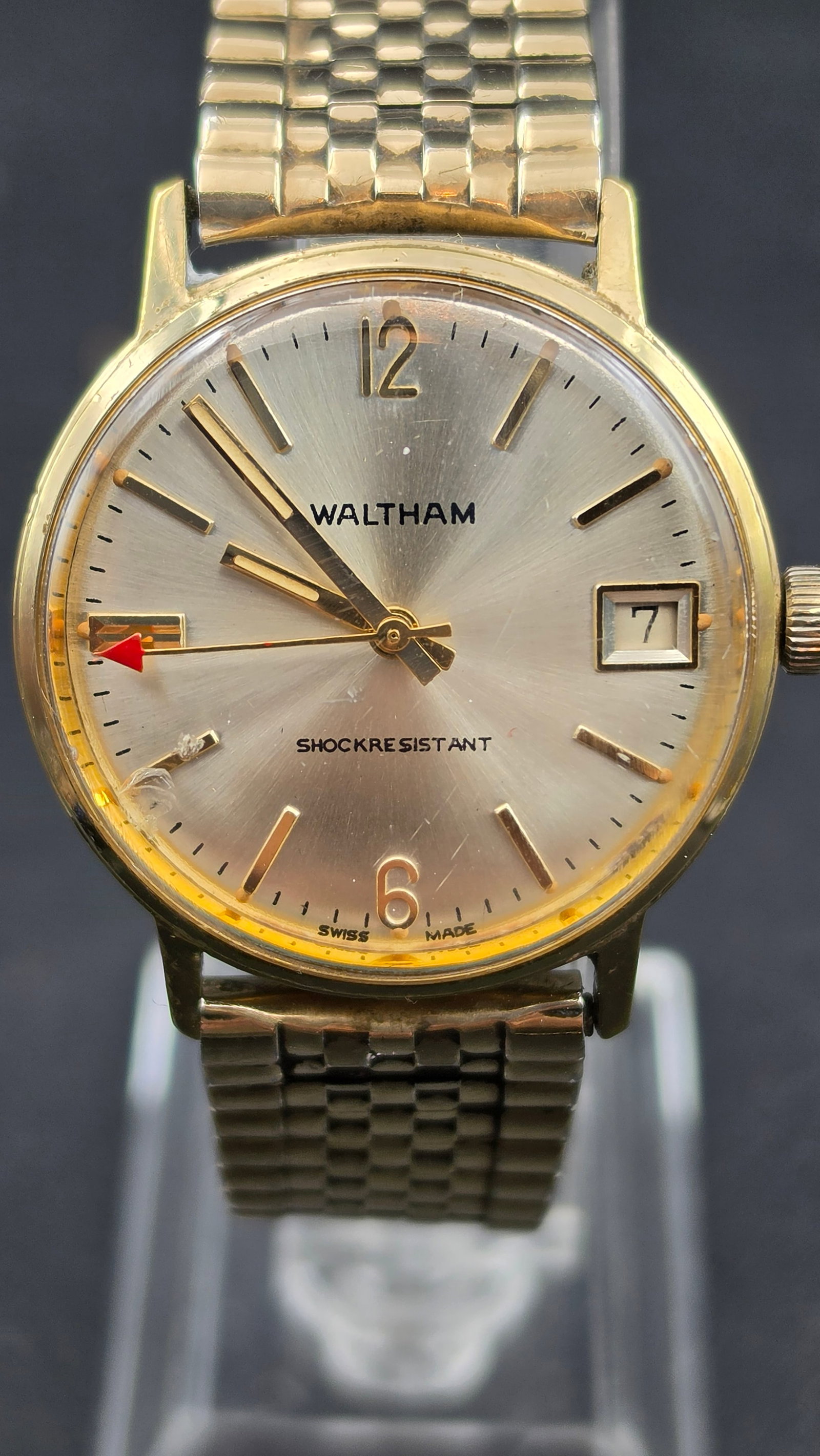 Vintage Waltham Watch Co Swiss 7 Jewel Mechanical Watch 32.80mm Shock Resistant Date Gold Tone: Champagne sunburst dial with Waltham text, applied gold-tone baton hour markers, Arabic numerals at 12 and 6, a red arrow indicator at 9 o'clock, date window at 3 showing 7, Shock Resistant text below