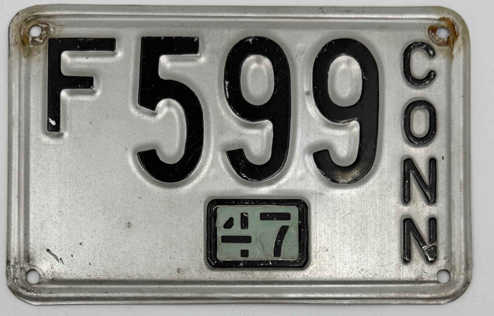 1957 Connecticut License Plate F599: Original 1957 Connecticut license plate with black embossed lettering on silver aluminum base and green “57” validation tab. Features vertical “CONN” lettering and plate number “F599