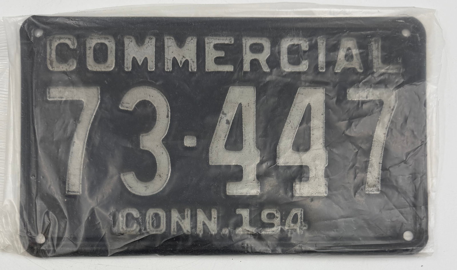 1934 Connecticut Commercial License Plate 73-447: Original 1934 Connecticut Commercial license plate, number 73-447. Black steel plate with white embossed lettering and “COMMERCIAL” across the top. Stored in protective plastic sleeve. Shows a