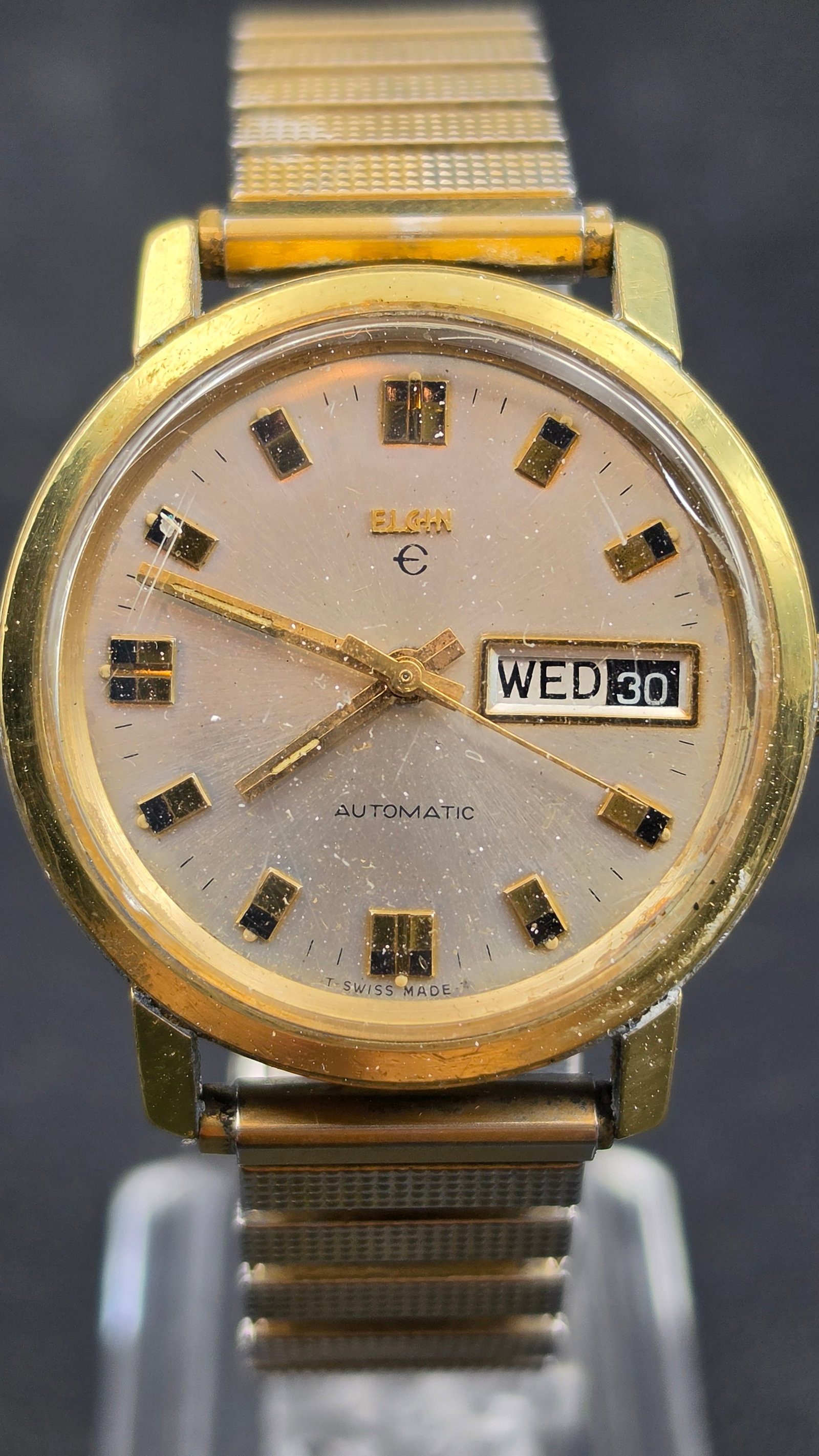 Vintage 1970s Elgin Automatic 17 Jewels Swiss Day-Date Watch Caliber 998 Gold Tone Case Runs: This is a vintage 1970s Elgin automatic wristwatch powered by the Swiss-made Elgin caliber 998, 17-jewel unadjusted self-winding movement visible through the exhibition caseback. The silver sunray dia