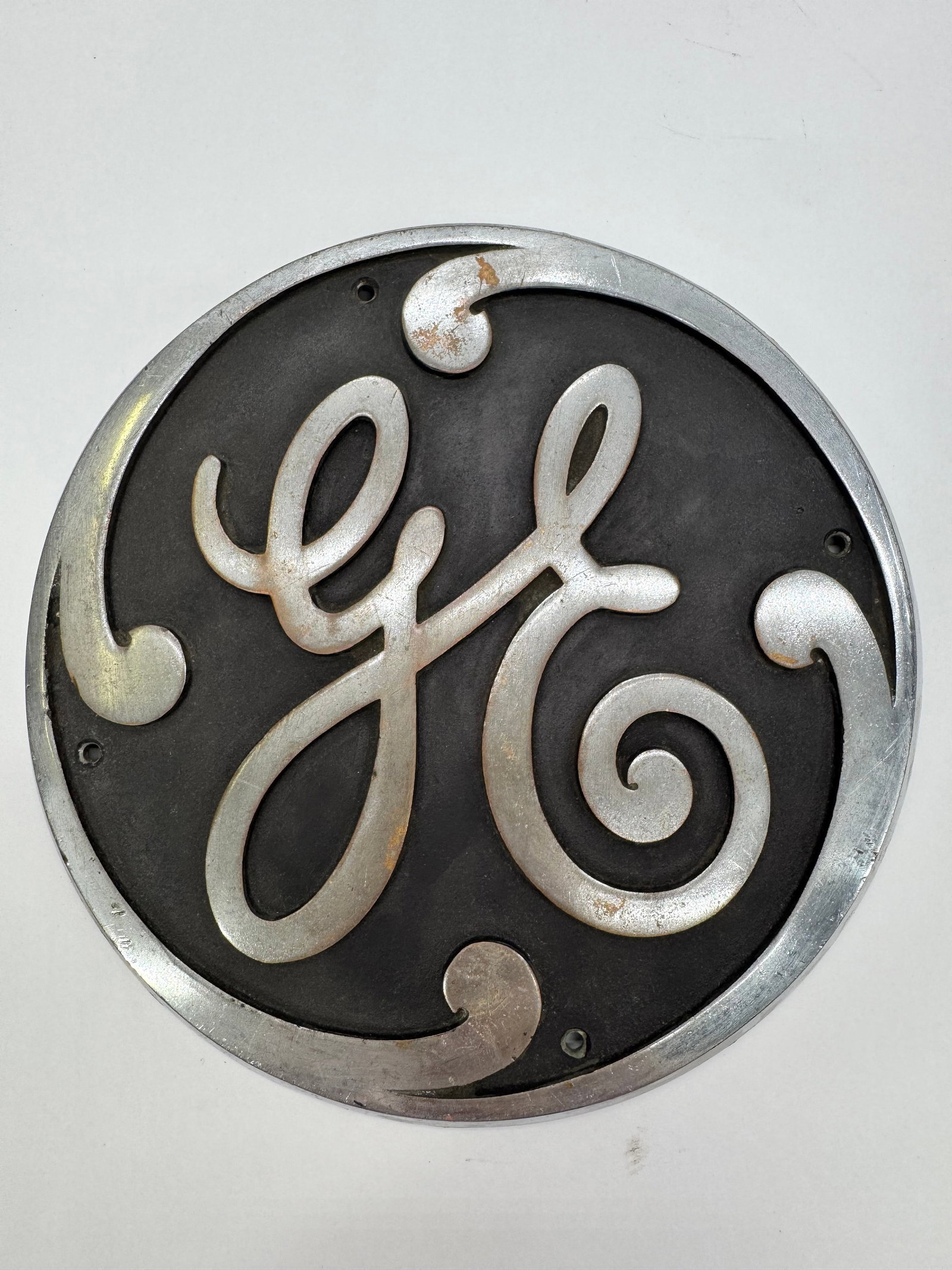 Vintage 8” General Electric GE Cast Metal Industrial Logo Sign Emblem: Original General Electric cast metal emblem featuring the iconic raised GE script logo. Measures approximately 8 inches in diameter and retains its original industrial finish with mounting holes intac