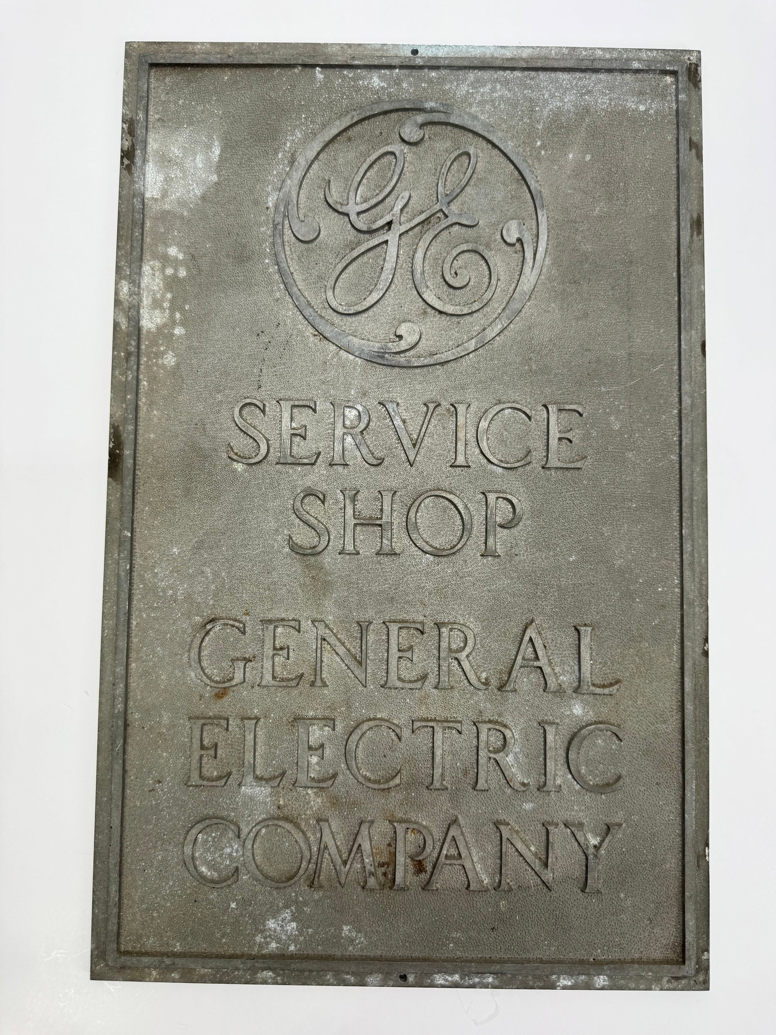 Vintage General Electric GE Service Shop Embossed Metal Sign: Original vintage General Electric Service Shop sign featuring the iconic GE script logo and embossed lettering. Constructed of cast aluminum/zinc with a textured background and mounting holes for disp