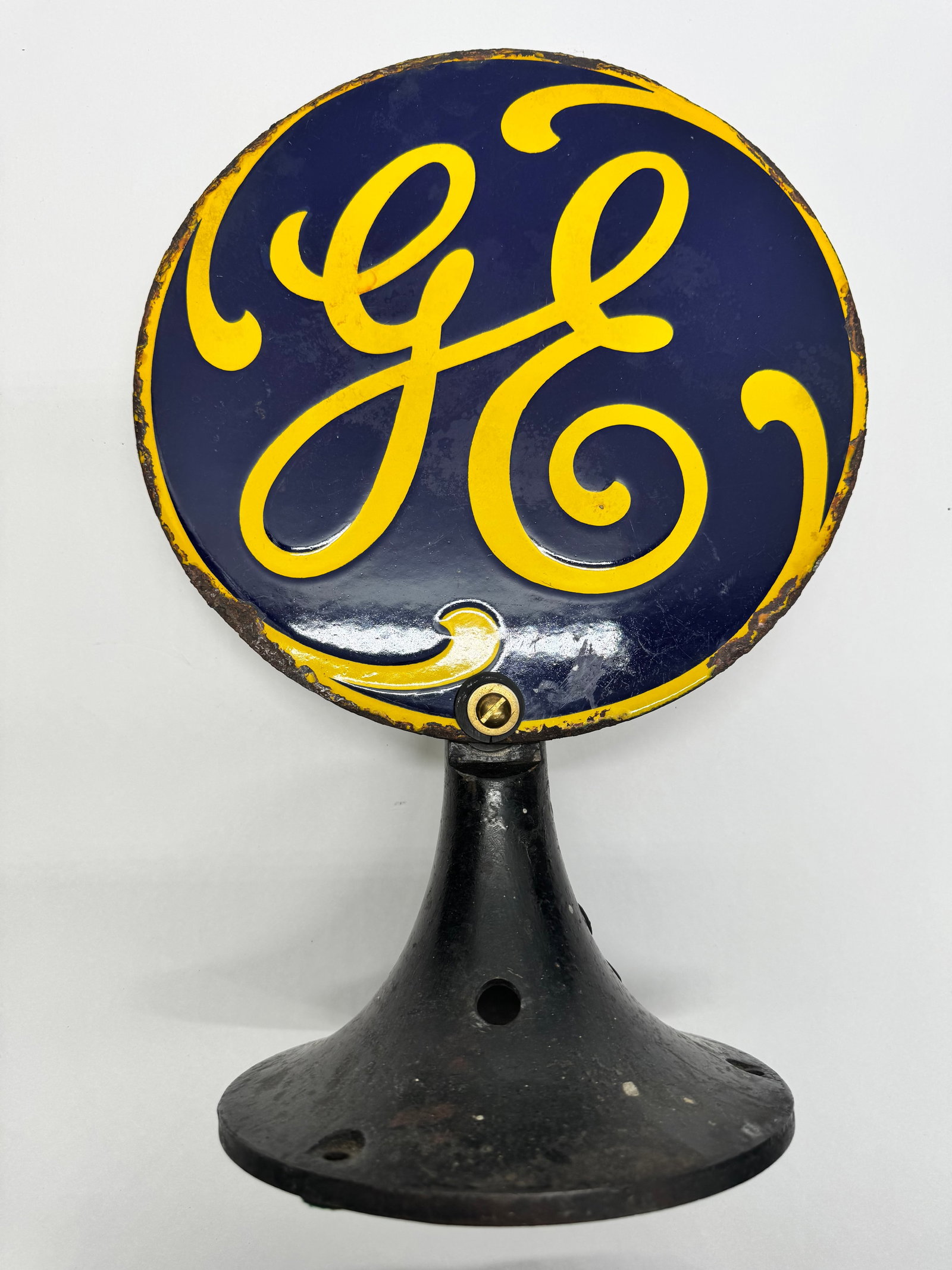 Vintage General Electric GE Porcelain Advertising Sign on Cast Iron Stand: Vintage General Electric porcelain enamel advertising sign featuring the iconic blue and yellow GE monogram mounted on a cast-iron pedestal base. Likely dating from the 1920s–1940s. Shows edge wear 