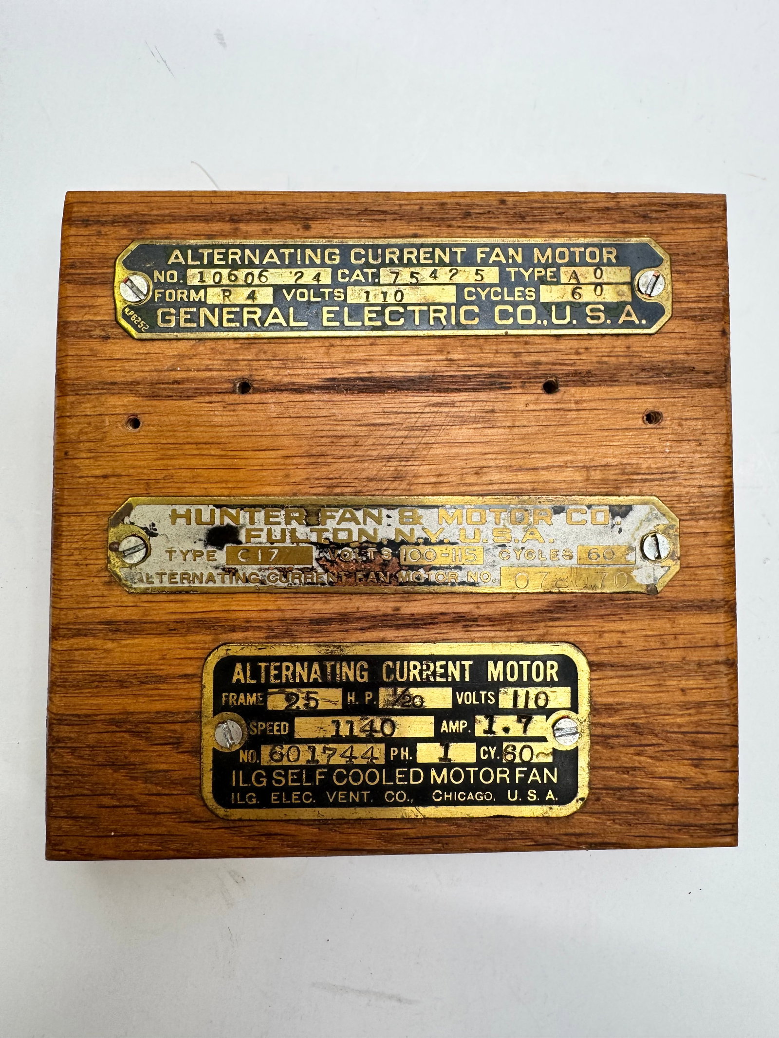 Vintage Electric Motor Brass Nameplate Display – GE, Hunter & ILG: Vintage display featuring three original brass electric motor nameplates mounted on a wooden base. Includes a General Electric Alternating Current Fan Motor plate (Model 10606), a Hunter Fan & Motor C