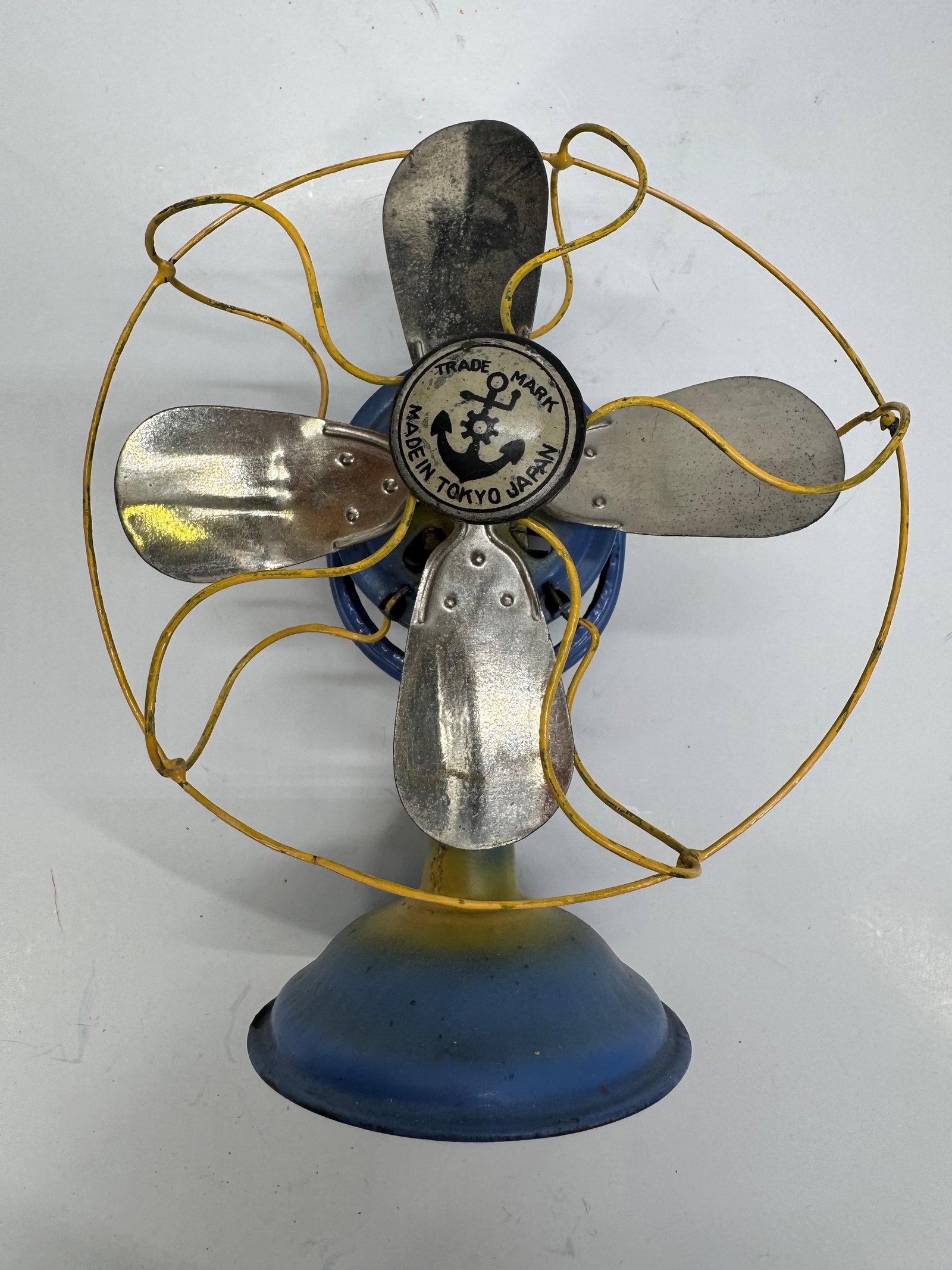 Vintage Tokyo Japan Tin Electric Desk Fan Anchor Logo: Vintage tin electric desk fan manufactured in Tokyo, Japan, featuring the anchor-and-gear trade mark on the blade hub. Finished in blue with a yellow wire safety cage and four metal blades. A charming