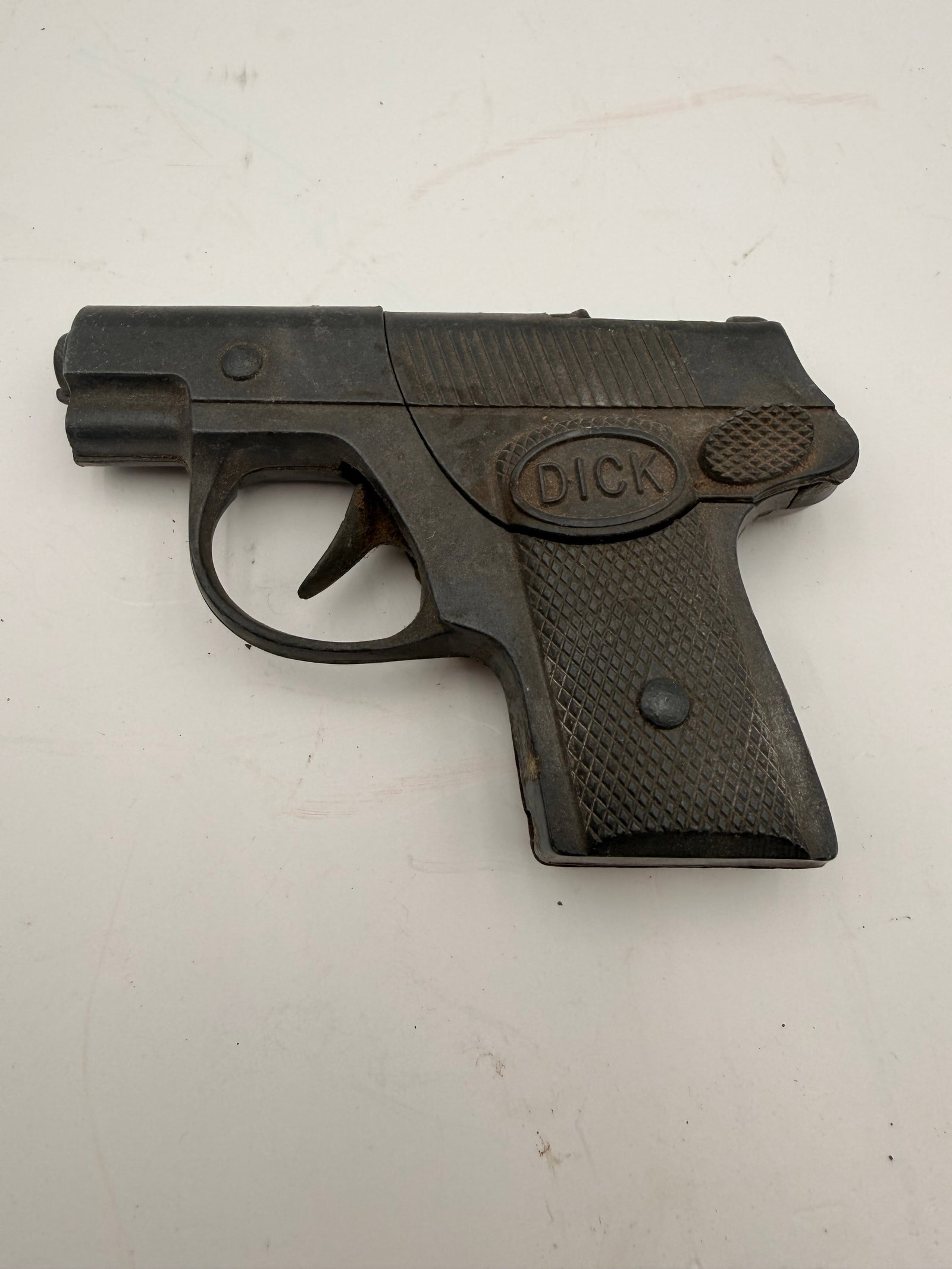 Vintage Cast Iron “DICK” Cap Pistol Toy: Vintage cast iron toy cap pistol with molded “DICK” grip panels and a compact semi-automatic pistol design. Features a dark painted finish, checkered grip, and cast metal construction. A