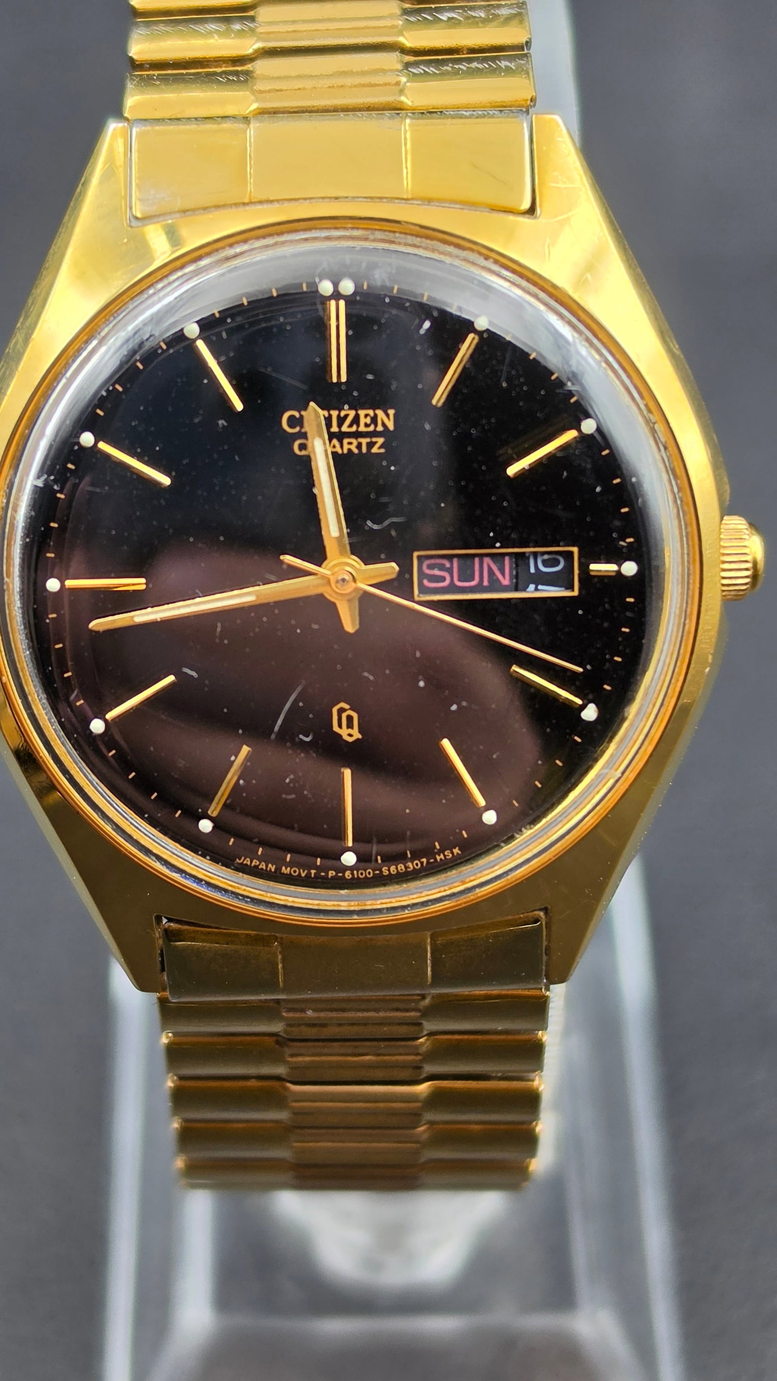 Citizen Quartz Day Date Gold Tone Dress Watch Cal 6100 CQ Logo Black Sunburst Dial Ref 6100-S68307: A vintage Citizen Quartz wristwatch in an all gold tone base metal case, case width measuring 36.13mm, with a smooth stepped bezel and an integrated gold tone bracelet. The black dial has a subtle sun