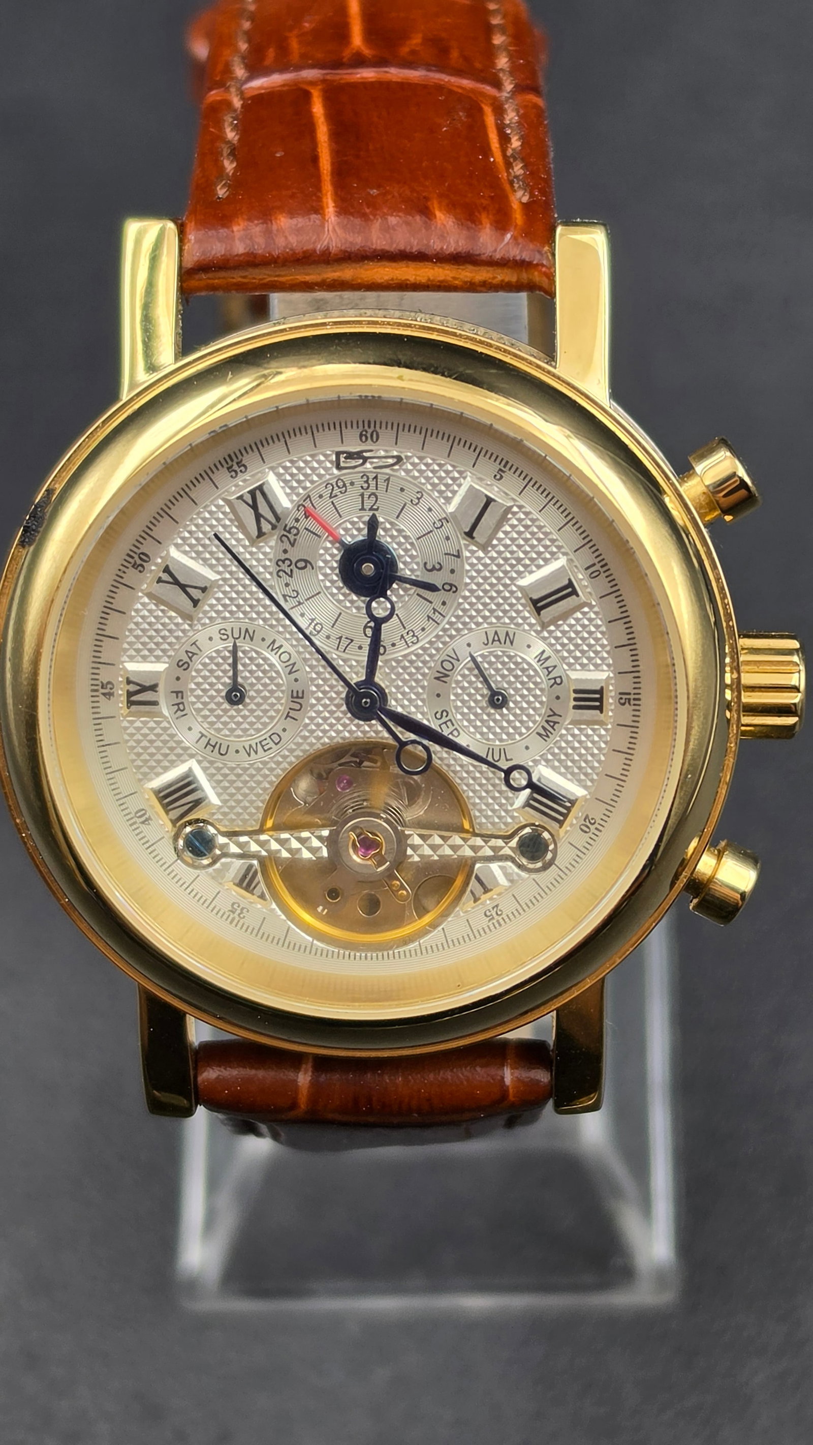 Daniel Steiger Automatic Watch Model DS 4056M Open Heart Day Month Date Gold Tone Case 35 Jewels: This is a Daniel Steiger automatic watch, model number DS 4056M. The silvered guilloche textured dial features an open heart cutout at 6 o'clock revealing the balance wheel, a day of the week subdial 
