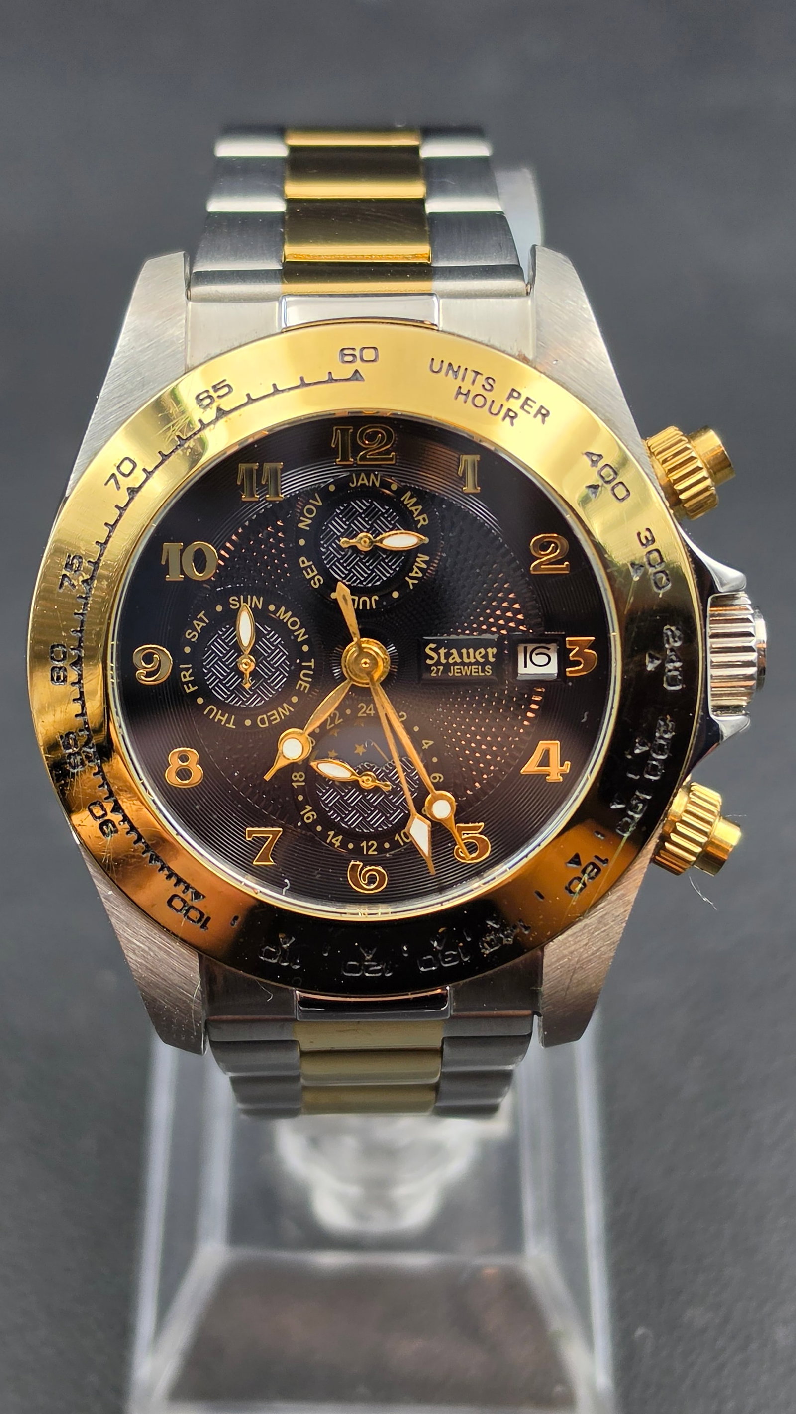 Stauer Automatic Watch 27 Jewels Reference 21222 Two Tone Day Month Moonphase Tachymeter: This is a Stauer automatic watch, reference 21222, with a multifunction dial. The black dial is signed Stauer 27 JEWELS and has gold tone Arabic numeral hour markers. Three sub-dials display the day o