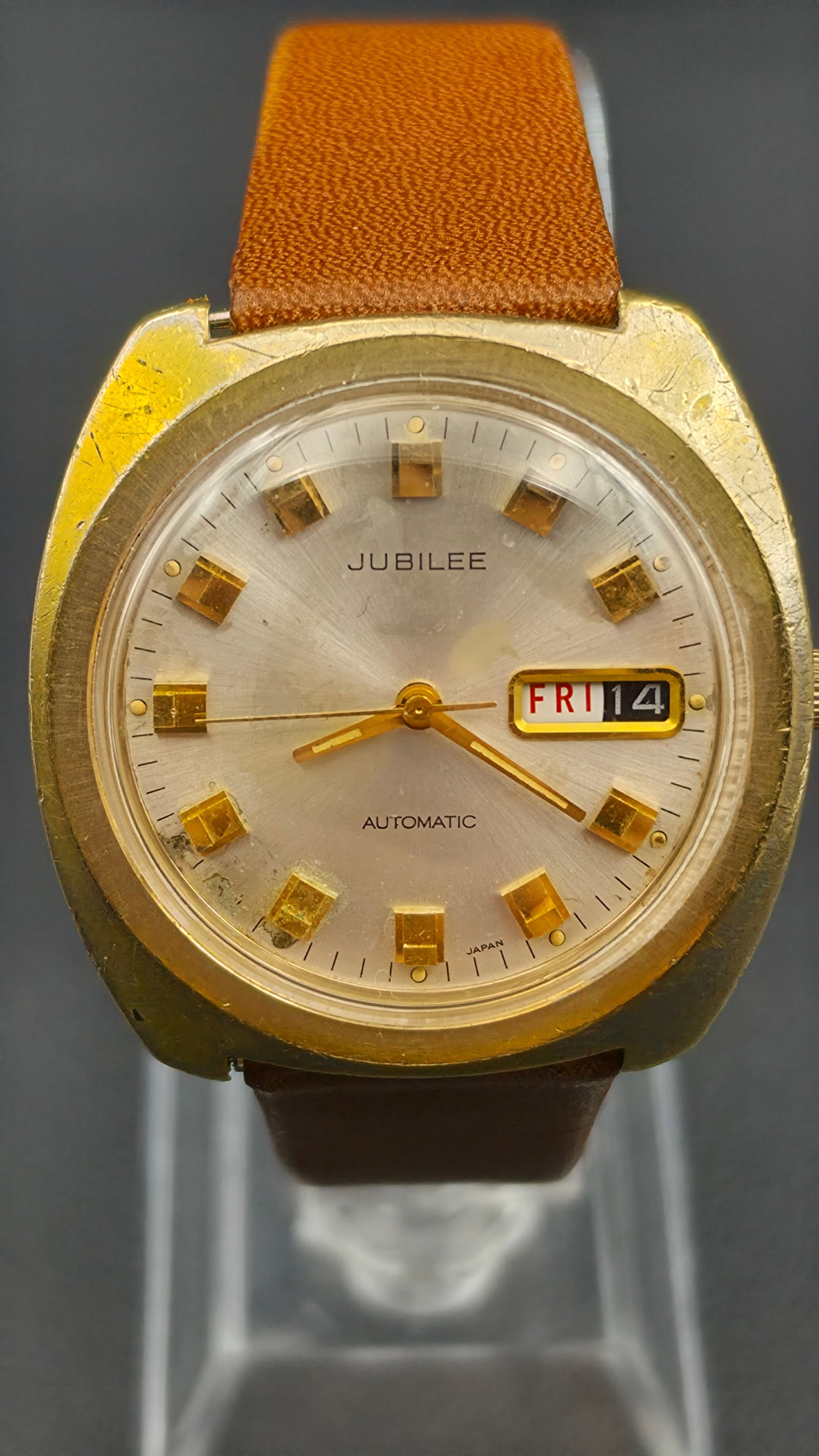 Vintage Jubilee Automatic 17 Jewel Day Date Gold Tone Cushion Case Mens Watch: Vintage Jubilee automatic day date wristwatch with a gold tone cushion shaped case measuring approximately 37.61mm across. Silver sunburst dial signed JUBILEE and AUTOMATIC with applied gold tone rect