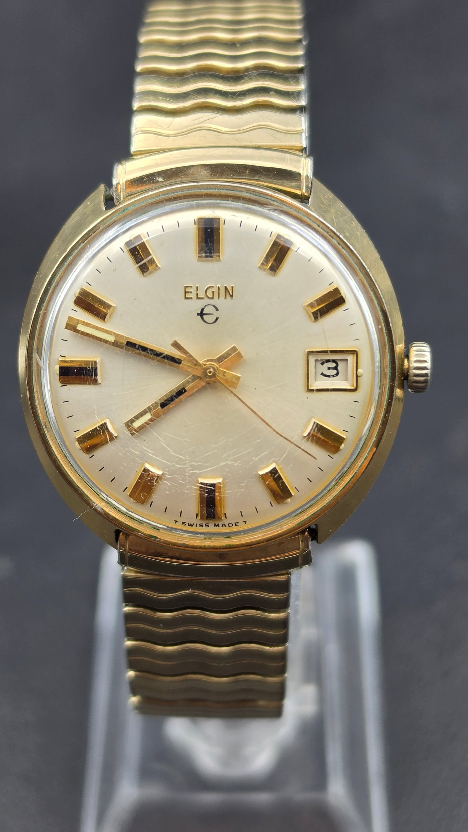 Elgin Swiss Made 1970s Mens Watch, Date Window, Base Metal Bezel: This is a 1970s Elgin men's wristwatch, Swiss made, in a round case with a base metal bezel and stainless steel back, measuring approximately 33.07mm wide excluding the crown. The dial is signed Elgin