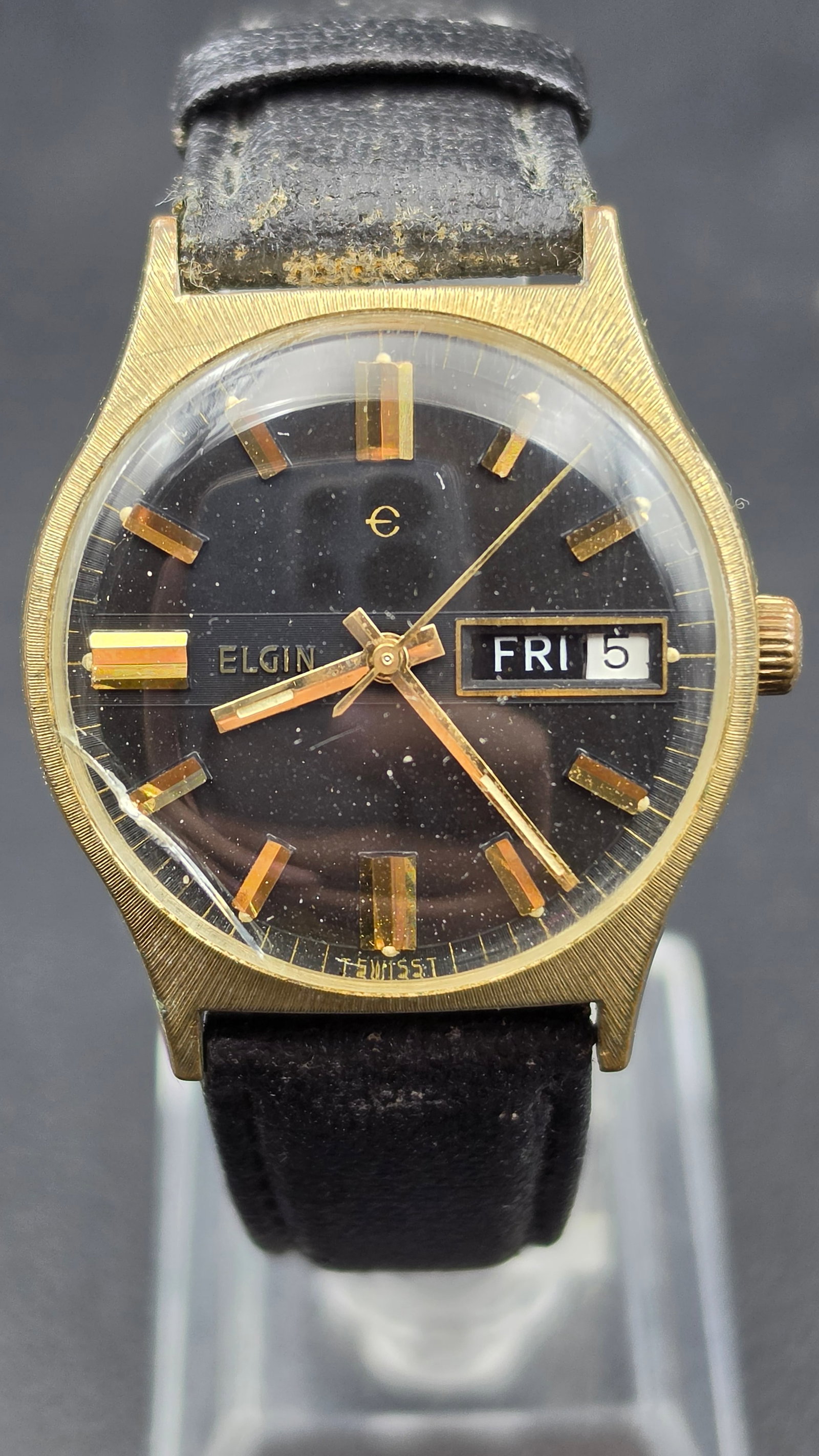 1970s Elgin Swiss Day Date Gold Tone Textured Case Watch: This vintage Elgin wristwatch has a gold tone case with a textured bark finish measuring approximately 33.34mm. The black dial is signed Elgin with the stylized "e" logo, applied gold baton markers, a