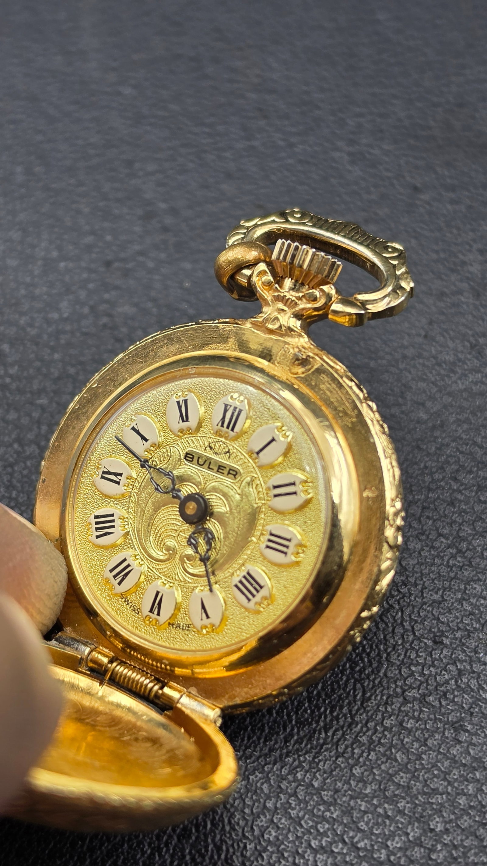 1960s Buler Swiss Gold-Tone Miniature Pendant Watch, Engraved Floral Case, Roman Numeral Cartouche D: This miniature pendant watch is signed Buler, Swiss Made on the gold-tone dial, with black Roman numerals set on individual white enamel cartouches and a scrolling engine-turned center. The case measu