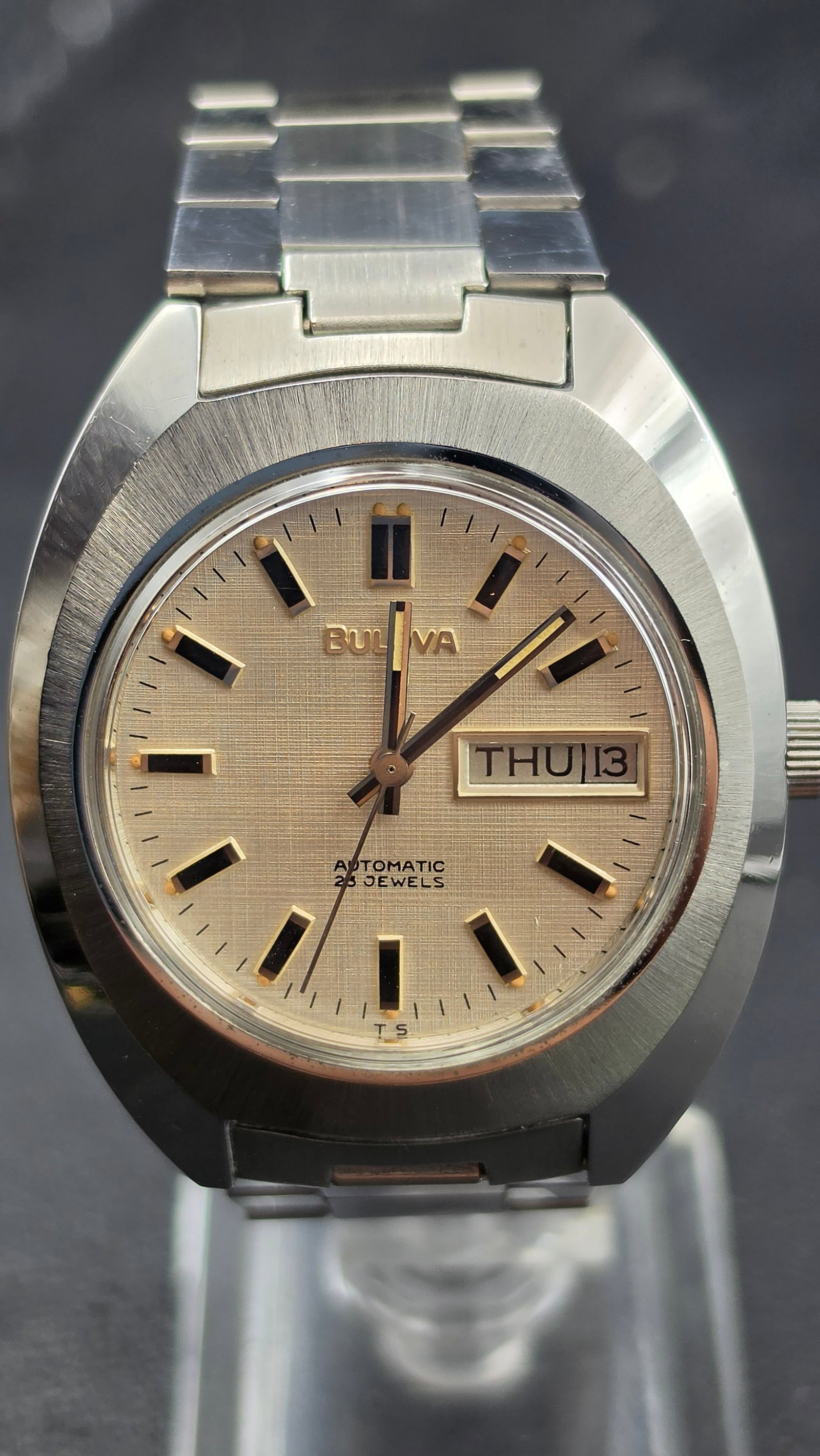 Bulova Automatic Day Date 23 Jewel Watch 1976 Running: This is a Bulova Automatic wristwatch with a cushion shaped stainless steel case. The dial is signed Bulova, Automatic, 23 Jewels, with a linen textured finish, raised baguette style hour markers, a d