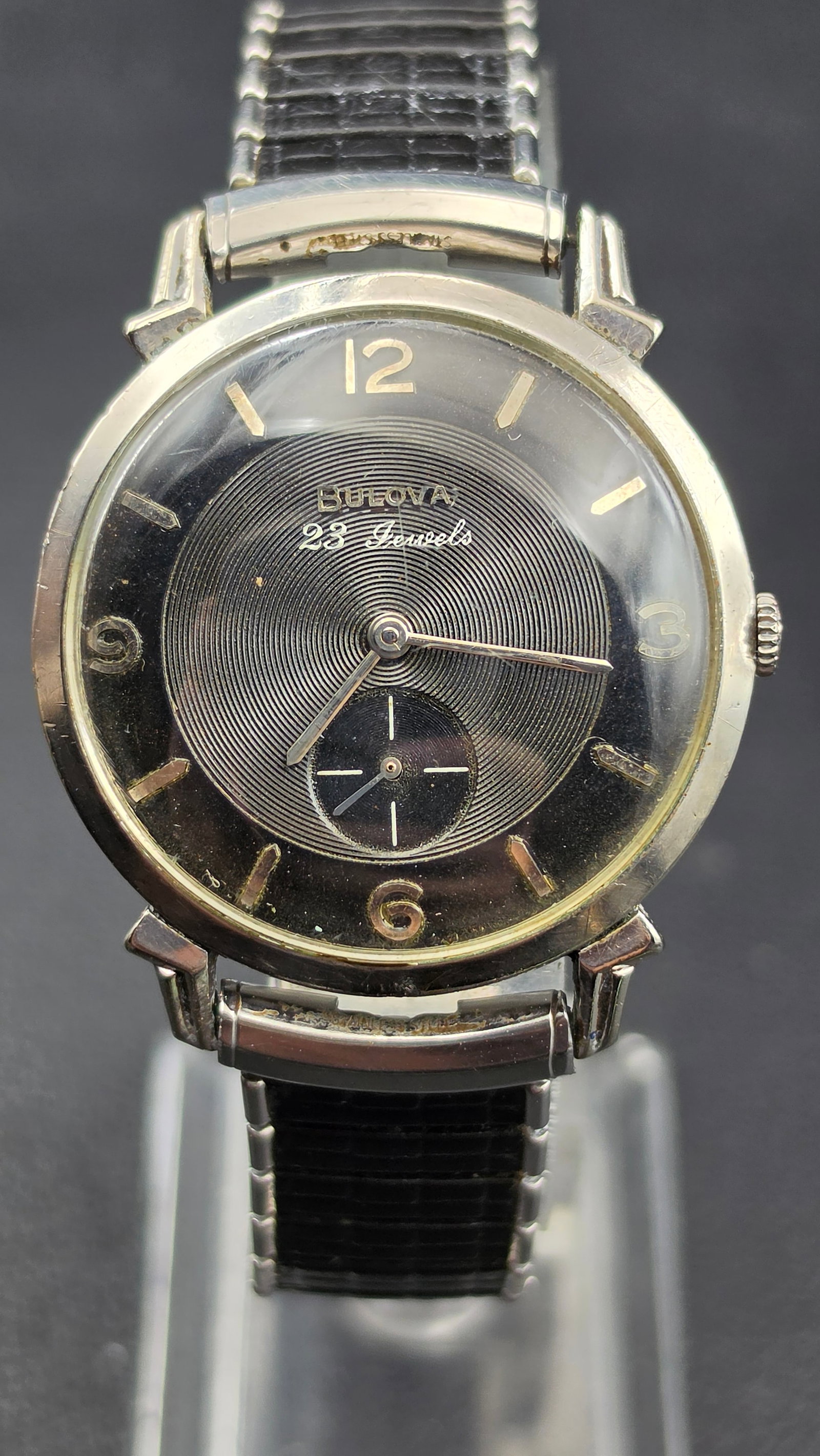 1958 Bulova 23 Jewel Cal. 10BP 10K RGP Men's Watch - Black Dial, 6 Adjustments: This vintage Bulova wristwatch features a black guilloche dial signed "Bulova 23 Jewels" with luminous arrow markers, Arabic numerals at 12, 3, 6, and 9, and a sub-seconds register at 6 o'clock. The c