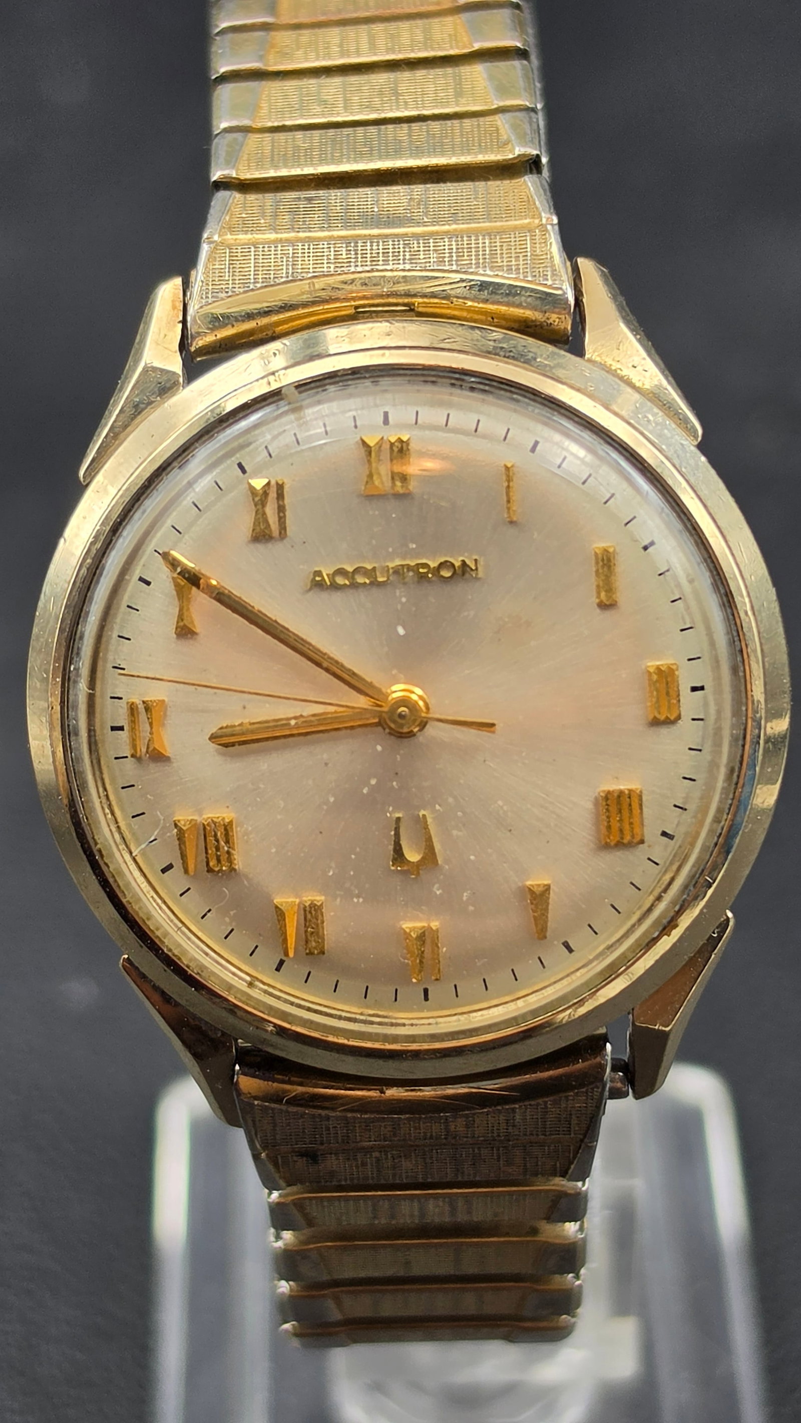 1970 Bulova Accutron 10K Gold Filled Watch, Roman Numeral Dial: This vintage Bulova Accutron wristwatch features a champagne dial signed "Accutron" with the tuning fork logo, applied gold-tone stylized Roman numeral markers, and dauphine hands. The dial shows visi