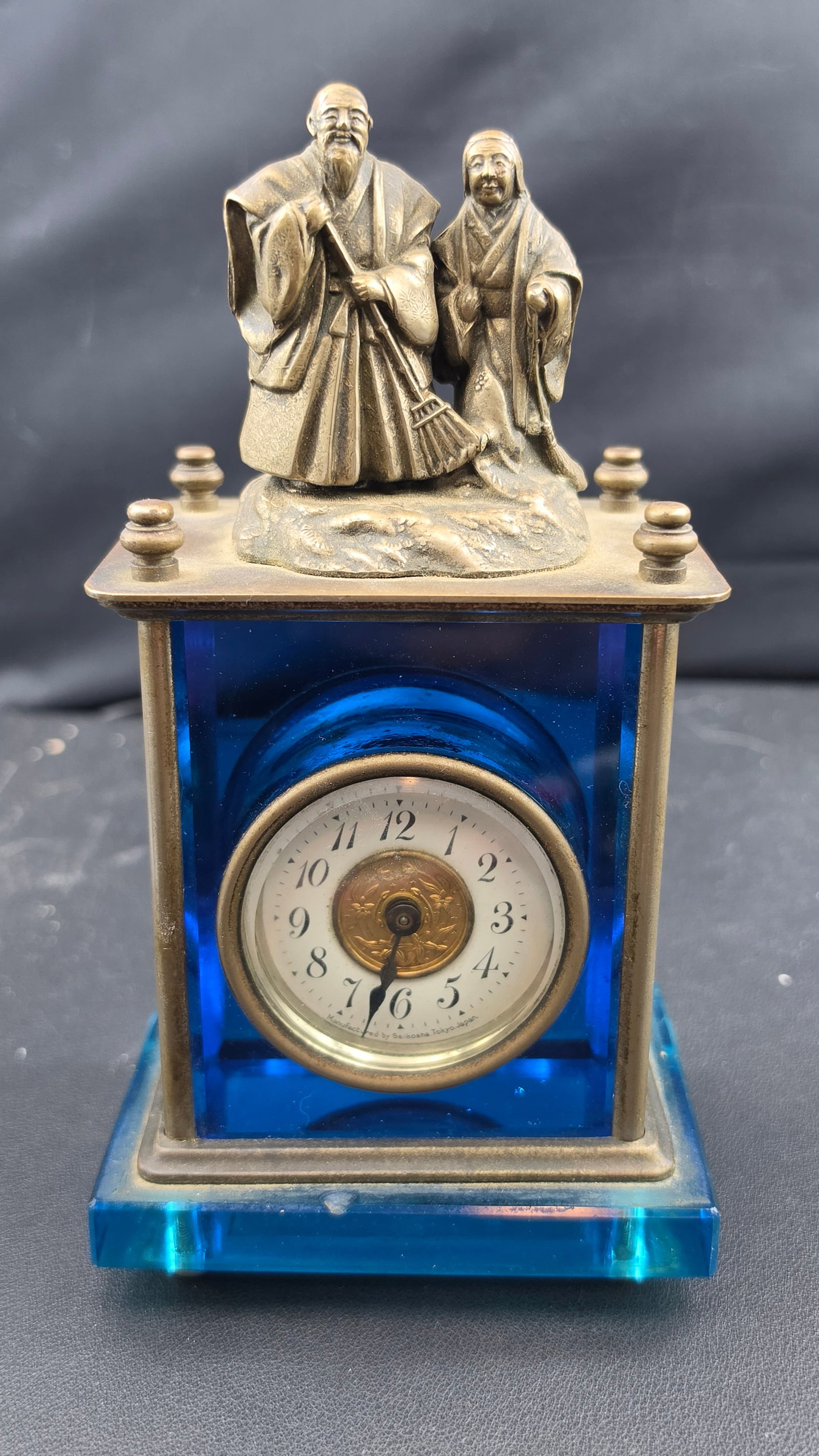 Seikosha Tokyo Japan Antique Bronze and Blue Glass Mantel Clock with Takasago Figures: This is an antique Seikosha Tokyo Japan mantel clock, its case constructed of cobalt blue glass panels held in a bronze frame, topped with a detailed bronze figural group depicting the Takasago couple