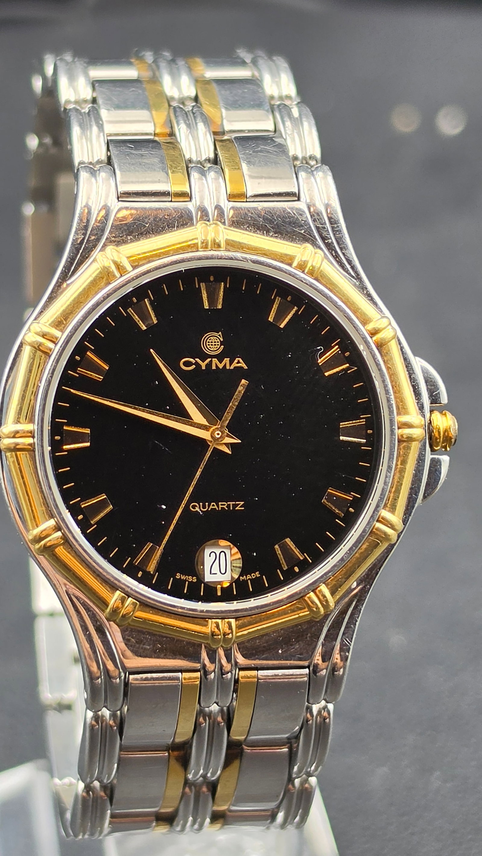 Cyma Le Locle Swiss Made Two Tone Quartz Watch 18K Gold Bezel: This is a Cyma Watch Ltd quartz watch from Le Locle, Switzerland, featuring a black dial with gold baton hour markers, a date window, and signed Cyma Quartz Swiss Made. The case back is stamped Stainl