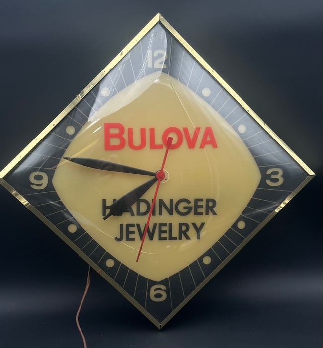 Vintage 1950s Bulova Lighted Advertising Clock – Hadinger Jewelry: Original mid-century Bulova lighted wall clock featuring red “Bulova” lettering and custom “Hadinger Jewelry” branding. Diamond-shaped acrylic face with black and cream dial and brass-