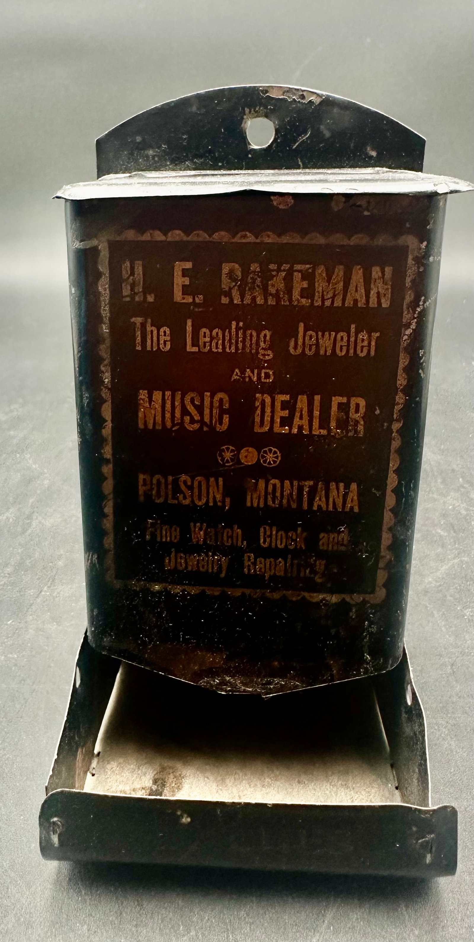 Antique H.E. Rakeman Polson Montana Advertising Match Holder: Antique tin wall-mounted match holder advertising H.E. Rakeman, jeweler and music dealer in Polson, Montana. Measures approximately 6” tall. Original lettering is faded, with scratches, paint lo