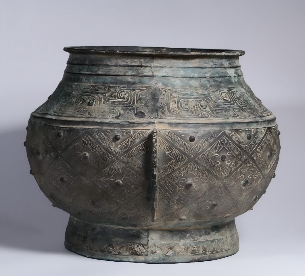A bronze geometric drum nail pattern urn from the Warring Han Dynasty: The jar is made of bronze and is preserved egg green. Round body, mouth edge outward, short neck, broad shoulders, bulging belly, circle feet. The neck is decorated with string patterns, and the shoul