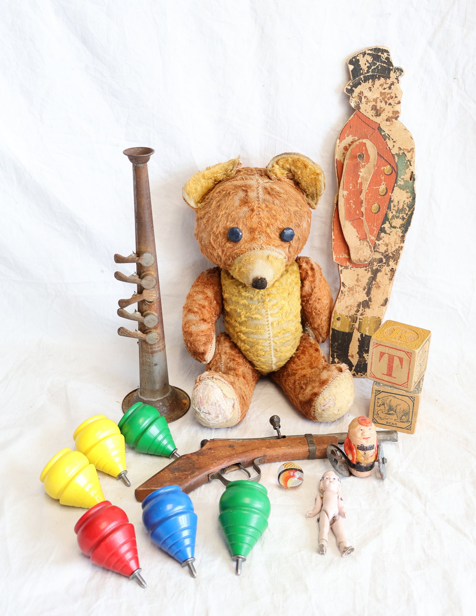 Lot of Children's Toys and Play Things; A Celluloid Rollie Pollie, Doll, Teddy, & Blocks: Rollie Pollie: 2 1/4" tall Teddy Bear: 13" Tin Horn, Bisque Doll, Blown Marble, 3 Blocks, and 6 Tops. Gurley Auction Company provides In House Packing and Shipping and strive to keep costs as reasonab