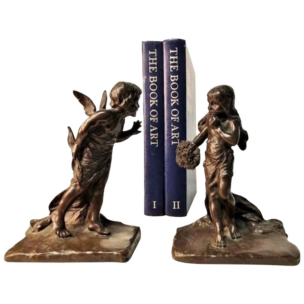 American Aestheticism, Griffoul Foundry Sculptural Bronze Bookends, Early XX C.: A charming set of bronze bookends. One of the bookends depicts a figure of a young girl with a questioning face expression, holding a bouquet; and the other a cupid, who peeks around the edge of the b