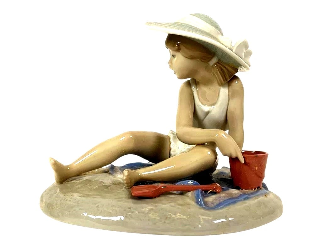 LLADRO RETIRED: 1993 SANDCASTLES: LLADRO RETIRED: 1993 SANDCASTLES Lladro porcelain figurine titled Playing in the Sand, No. 5488. Depicts a young girl seated on a textured sandy base, wearing a white swimsuit and wide-brimmed hat, ho