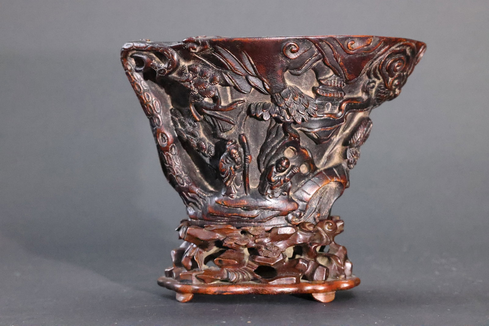 An Aloeswood 'Landscape and Figures' 'Rhinoceros Horn-Imitation' Libation Cup, Qing Dynasty: Estimated Date: Mid- to Late Qing Dynasty. A carved agarwood (chenxiang) libation cup with landscape and scholar decoration, accompanied by its original hardwood stand.Attribution: This finely carved 