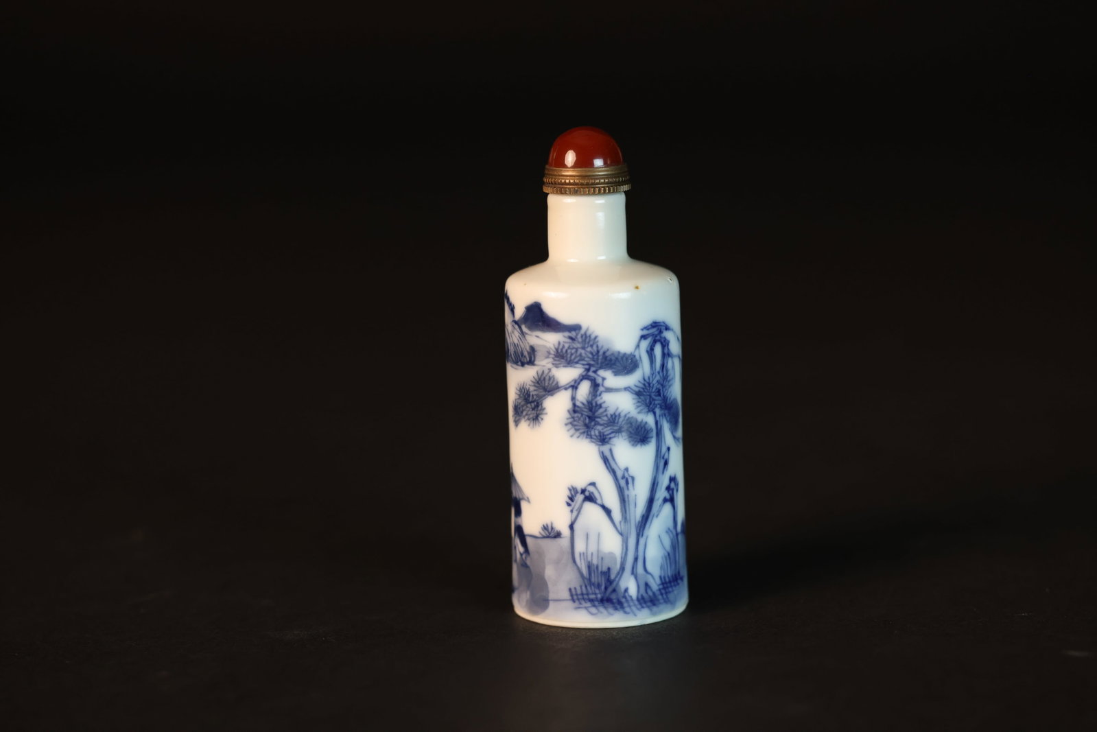 Qing Dynasty Blue-and-White Figural Snuff Bottle with Pine-and-Rock Motif, Agate Finial: Naturally aged with one minor glaze flake about the size of half a sesame seed on the body. The footrim has two similar small glaze flakes. Otherwise in excellent condition. H 9.1cm W 3.2cm