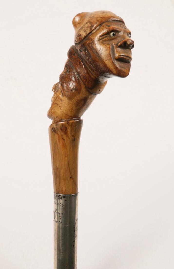 FOLK ART CANE: Maple Root Handled Oak Cane having a carving of two heads, stacked and facing in opposite directions, the uppermost with a knit cap, both having glass eyes, a broad silver band below, engraved "Lorenz
