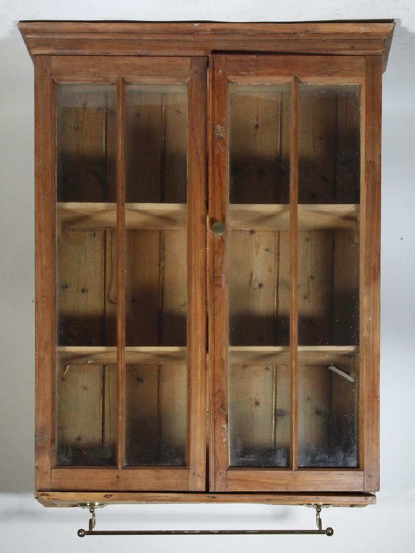 SMALL HANGING CUPBOARD: Irish Stripped Pine Wall Cupboard with molded cornice, two glass doors over three shelves, later underhung brass towel rack, 34" x 25" x 10". Good condition.
