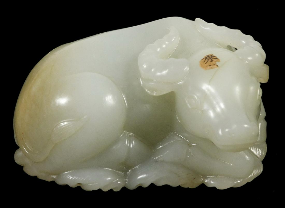 19TH C. CARVED CHINESE JADE FIGURINE: Celadon Jade Recumbent Water Buffalo, resting on an oval pad of water symbol, touches of russet on rump and one ear, remnant of an early paper label on his head, 2 5/8" long. Fine condition.