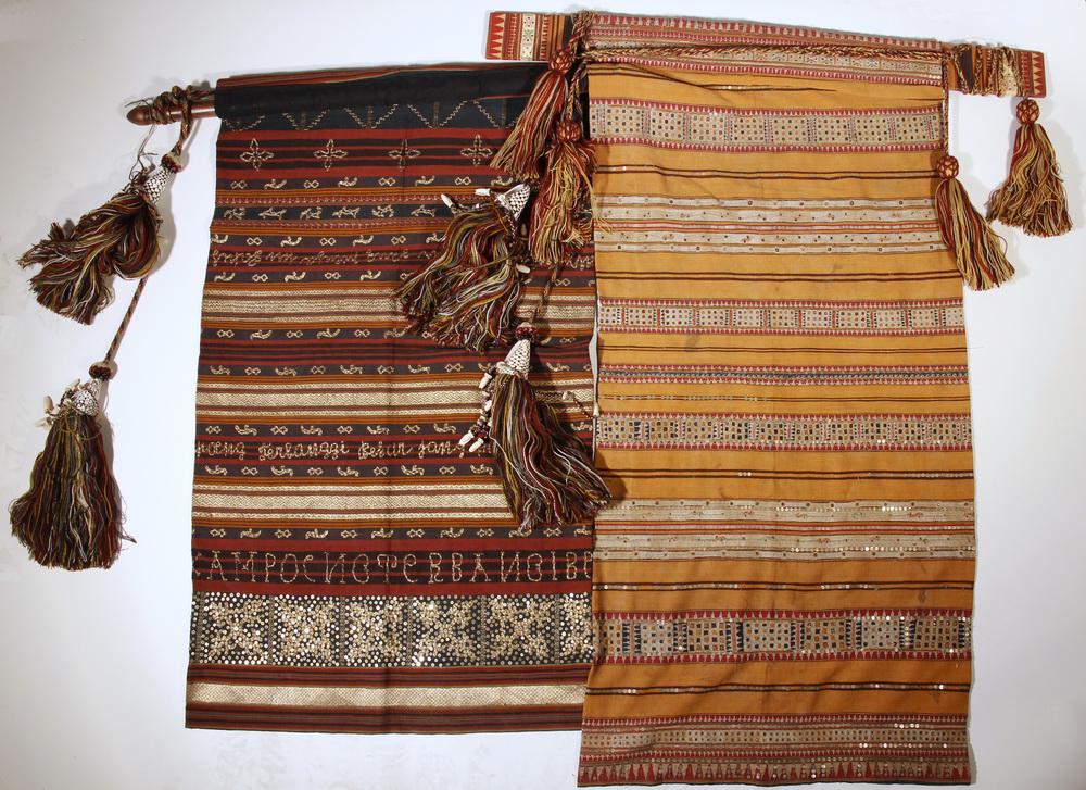 (2) INDONESIAN HANGING TEXTILES: Lot of (2) Indonesian Woven Hanging Textiles with tassels and hangers, including: Striped banner with metallic embroidery, embroidered maker's name 'Sara Dana Basaraia', on carved wooden hanger, flank