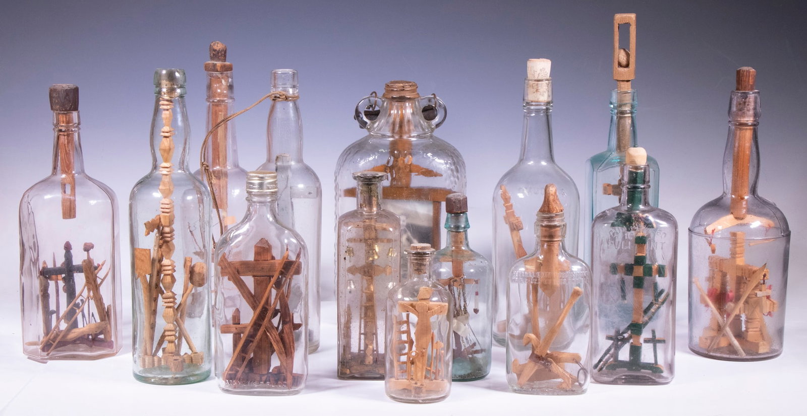 (14) AMERICAN FOLK ART WHIMSEYS IN BOTTLES: Vintage 1900-1930, of carved wood, some painted, within bottles of various origin, 6" to 13 1/2" tall, consisting of (10) Religious, (3) Marine & (1) in a grape jug, housing a photo of a WWI US Doughb