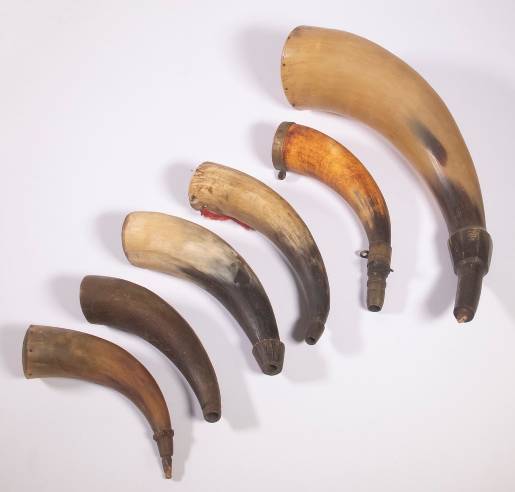 (6) POWDER HORNS: A collection of assorted powder horns. One utilizes brass fittings with a spring loaded latch and nozzle while the other five rely on simply wooden plugs at the tip of the horn. A couple of the horns 
