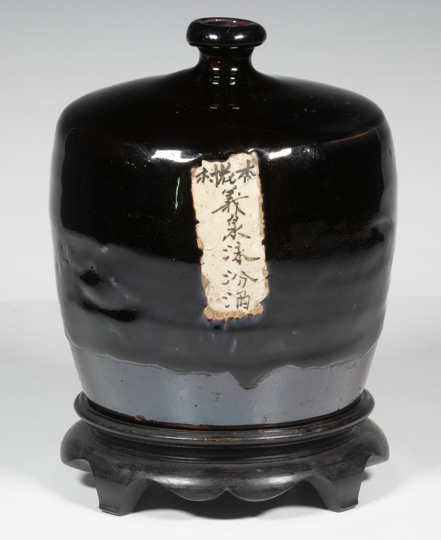 CHINESE STONEWARE WINE JAR ON STAND, 19TH CENTURY: 19th c. Black Glazed Wine Pot, with affixed paper Chinese character label, with carved wooden stand. Converted dimensions: 7 x 6 x 6 in. (17.8 x 15.2 x 15.2 cm.)