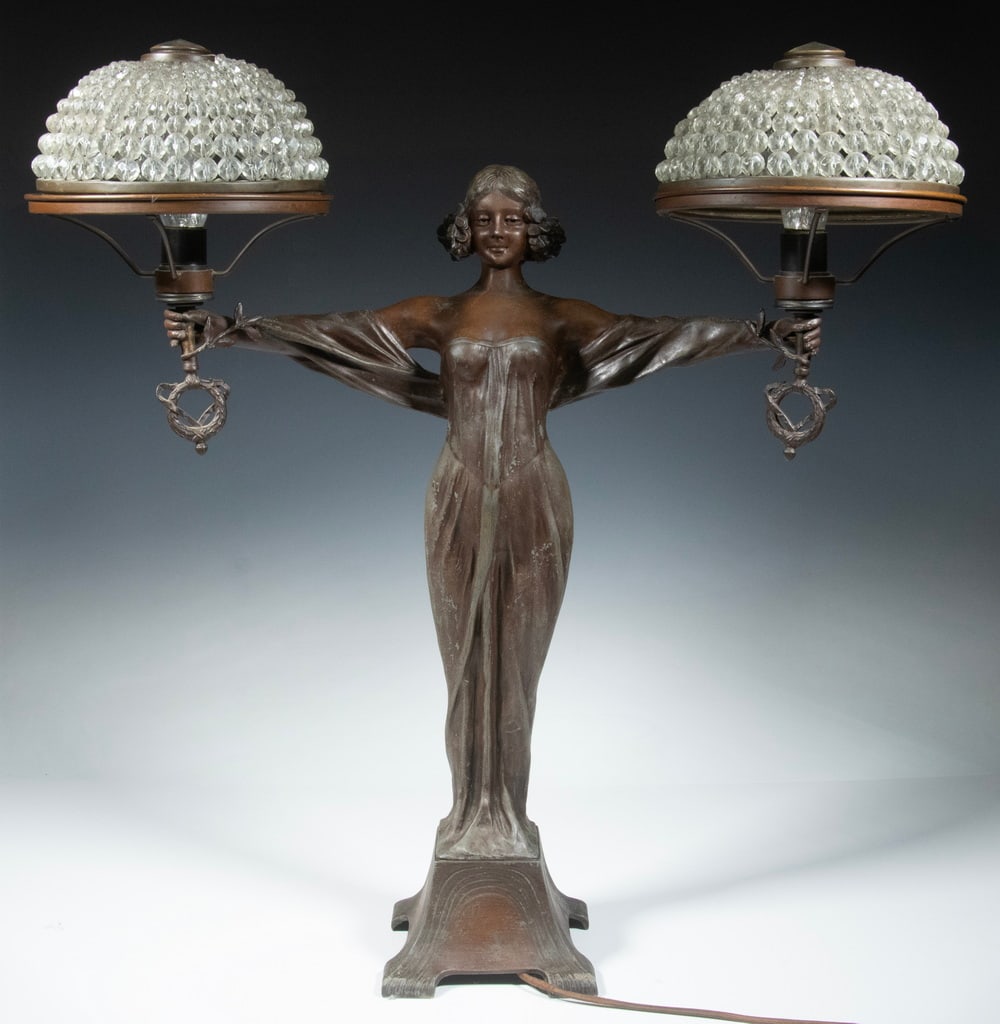 ART NOUVEAU LAMP AFTER SAINT-GAUDENS: Woman Holding Two Torches Aloft, bronze finished spelter lamp with domed beaded glass shades, having appropriate cloth covered cord and in-line switch, roughly 26" x 24" x 8", wear to finish. Converte