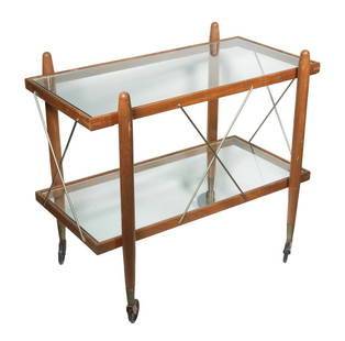 AMERICAN DANISH MODERN STYLE BAR CART: Teak Framed Two Glass Shelf Cart with round double tapered corner posts, brass X-frame on each side, resting on brass and black plastic wheels, circa 1960s.From the estate of Brooke Astor at her summe