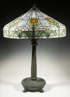 TALL ARTS & CRAFTS ERA BRONZE TABLE LAMP WITH TULIP SHADE: Four footed bronze base with incised Art Nouveau blossoms, with a new-classical tapered fluted column, flat sided tapered leaded shade of polychrome tulips alternating with sky blue panels. Unmarked,