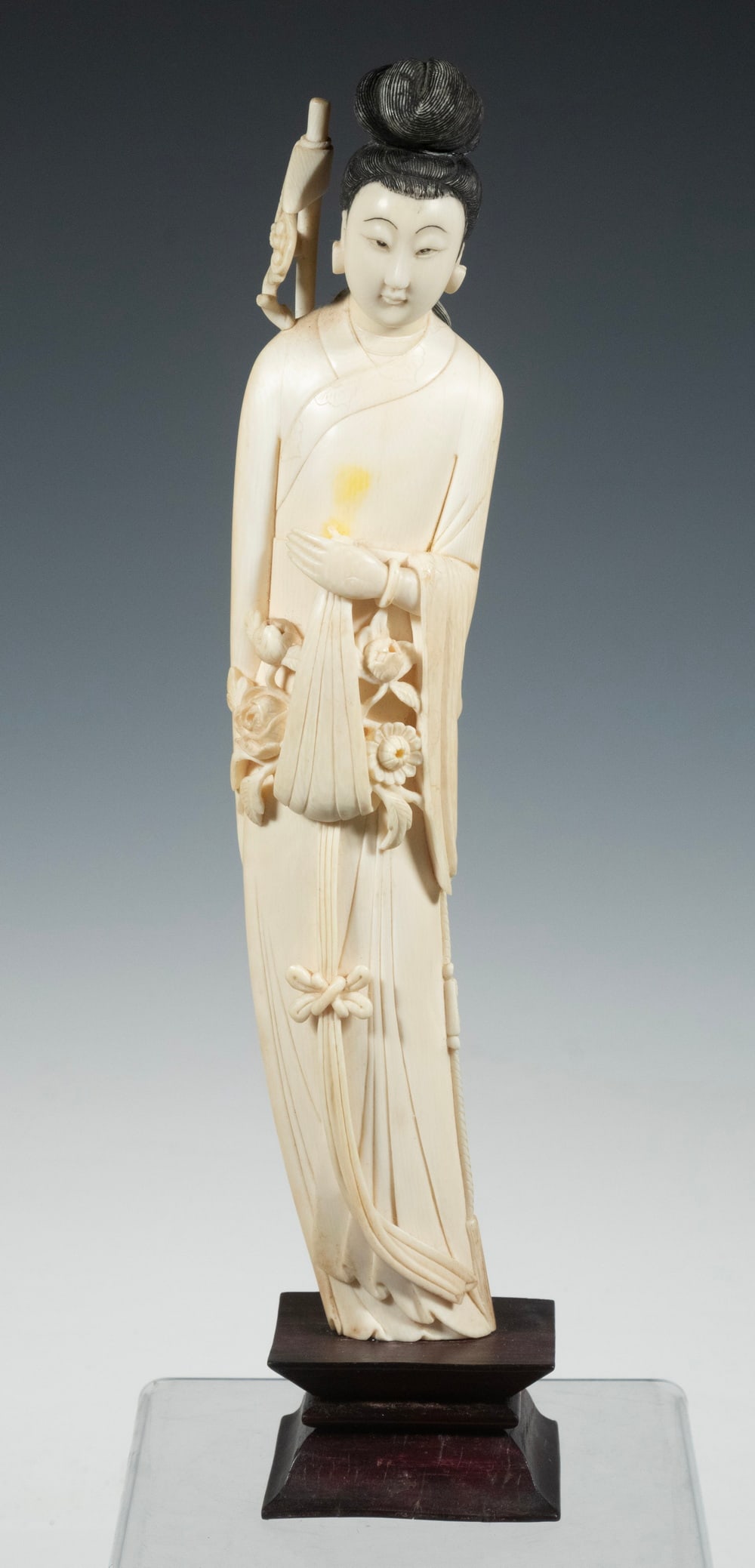 19TH C. JAPANESE IVORY OKIMONO: Graceful Standing Geisha, with lingzhe staff, flowers bundled in her robe, supported by her left hand, on wooden plinth. Height w/ Plinth: 14 1/2 in. (36.8 cm.)