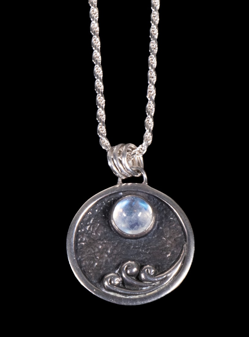 ITALIAN STERLING SILVER AND MOONSTONE PENDANT NECKLACE: An Italian sterling silver necklace featuring a circular pendant set with a round cabochon moonstone, exhibiting blue adularescence. The pendant is decorated with a stylized wave motif against a darke