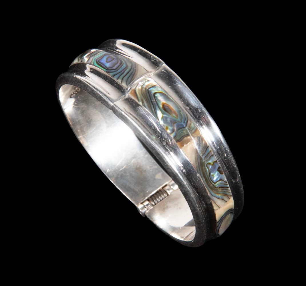 MEXICAN STERLING SILVER AND ABALONE HINGED BRACELET: A wide sterling silver hinged cuff bracelet inlaid with iridescent abalone shell panels, exhibiting vibrant green and violet hues. Marked “Mexico 925.” Inner circumference approx. 6 3/4 in