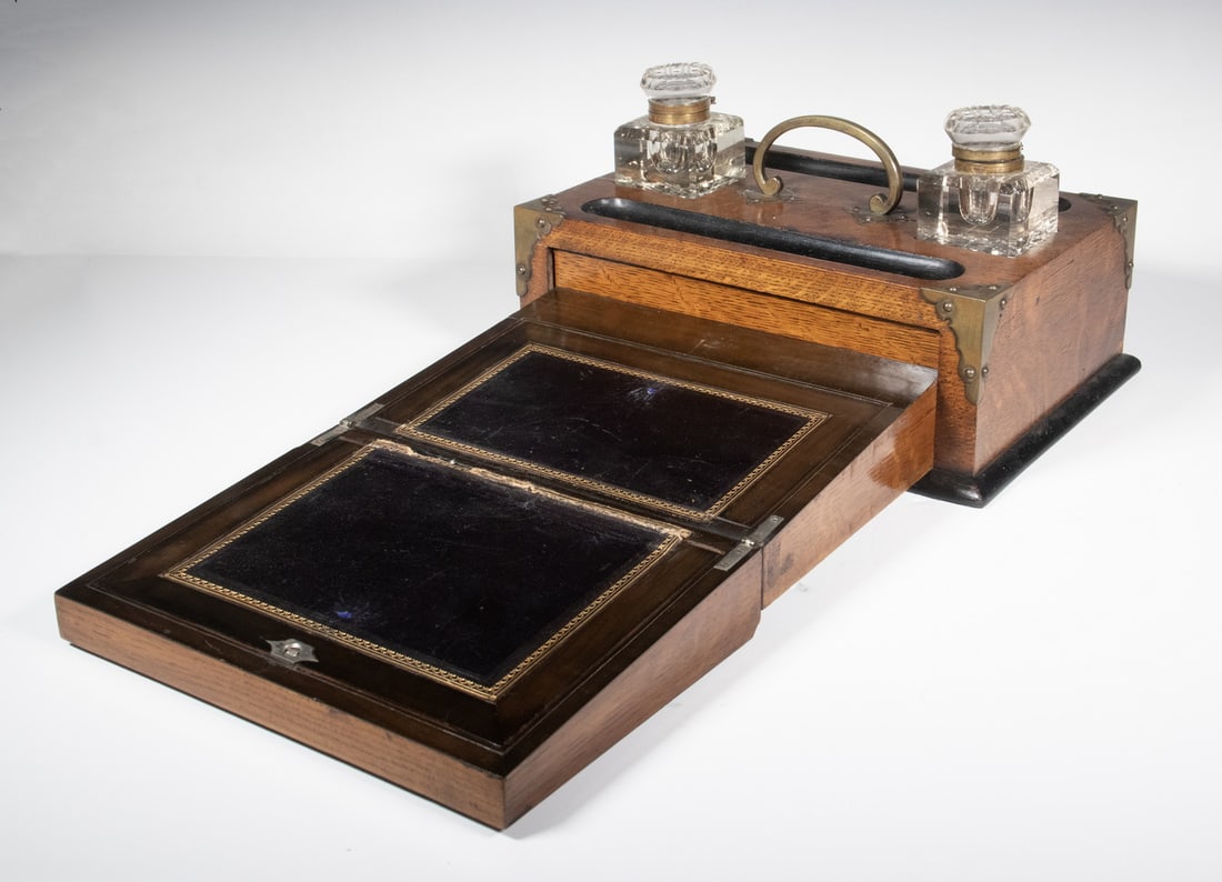 VICTORIAN OAK AND BRASS DOUBLE INKWELL WRITING SLOPE: A Victorian oak writing slope fitted with two cut-glass inkwells, brass mounts and carrying handle, and a hinged front opening to a leather-lined writing surface with gilt tooling. The top features re