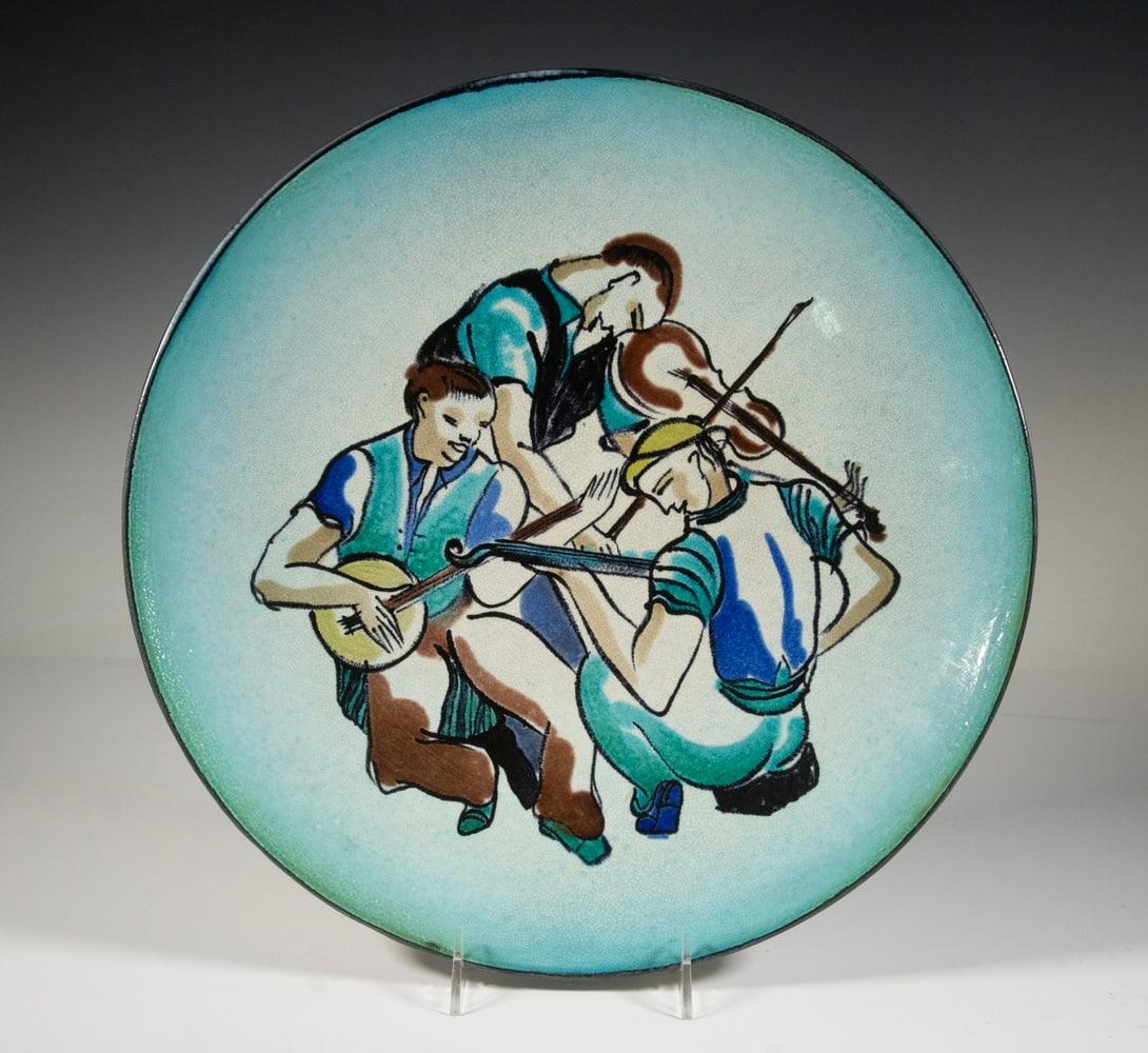 WHITNEY ATCHLEY GLAZED POTTERY WALL PLATE WITH THREE MUSICIANS, DATED 1937: A circular pottery wall plate featuring a vibrant hand-painted depiction of three musicians in blue-toned garments, playing banjo and two fiddles. Signed and dated “Whitney Atchley '37”