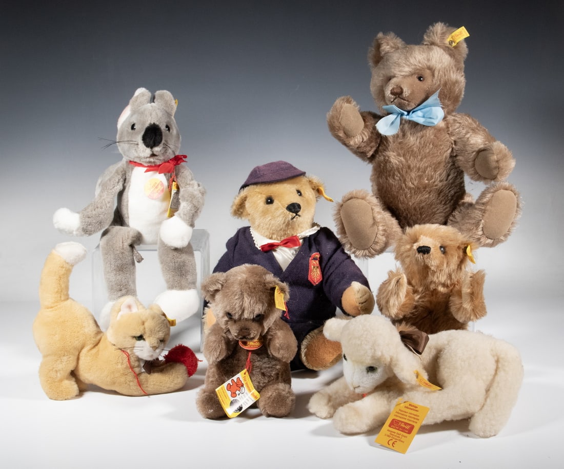 (7) STEIFF PLUSH ANIMALS WITH BUTTON-IN-EAR TAGS: A group of seven Steiff plush animals, each retaining the signature “button in ear” tag. Includes a classic jointed growler bear with blue ribbon; a dressed bear in navy outfit and cap; a