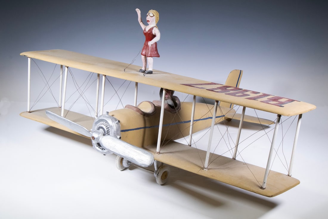 FOLK ART BIPLANE WITH PILOT & WING WALKER: Late 20th c. Biplane in carved and painted wood, marked "B1987", having a woman in a red dress wing walking, the moustachied pilot raising his hand to indicate he is going to land. Unsigned.16 1/2 x