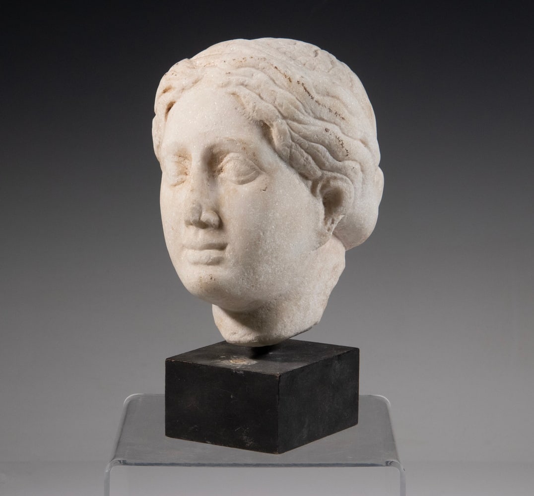 ROMAN MARBLE HEAD OF WOMAN: Fragment Head of Young Woman in white marble, having coiffed hair and a smile, on black block base.Height With Base: 9 in. (22.9 cm.), Head Only : 5 x 5 in. (12.7 x 12.7 cm.)