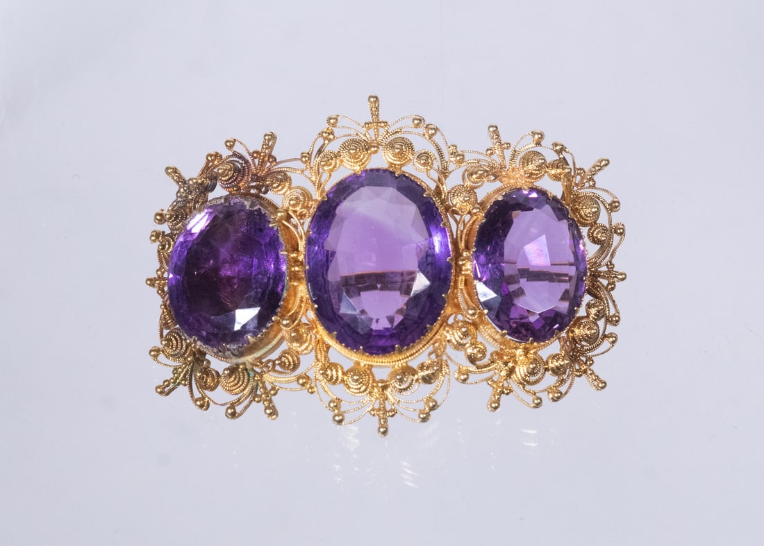 GEORGIAN CANNETILLE 18K GOLD AND AMETHYST BROOCH, EARLY 19TH CENTURY: A Georgian-era brooch set with (3) oval-cut purple amethysts, largest amethyst measuring 19mm X 15mm, mounted in tested 18K yellow gold. The stones are arranged in a horizontal trio within an elaborat